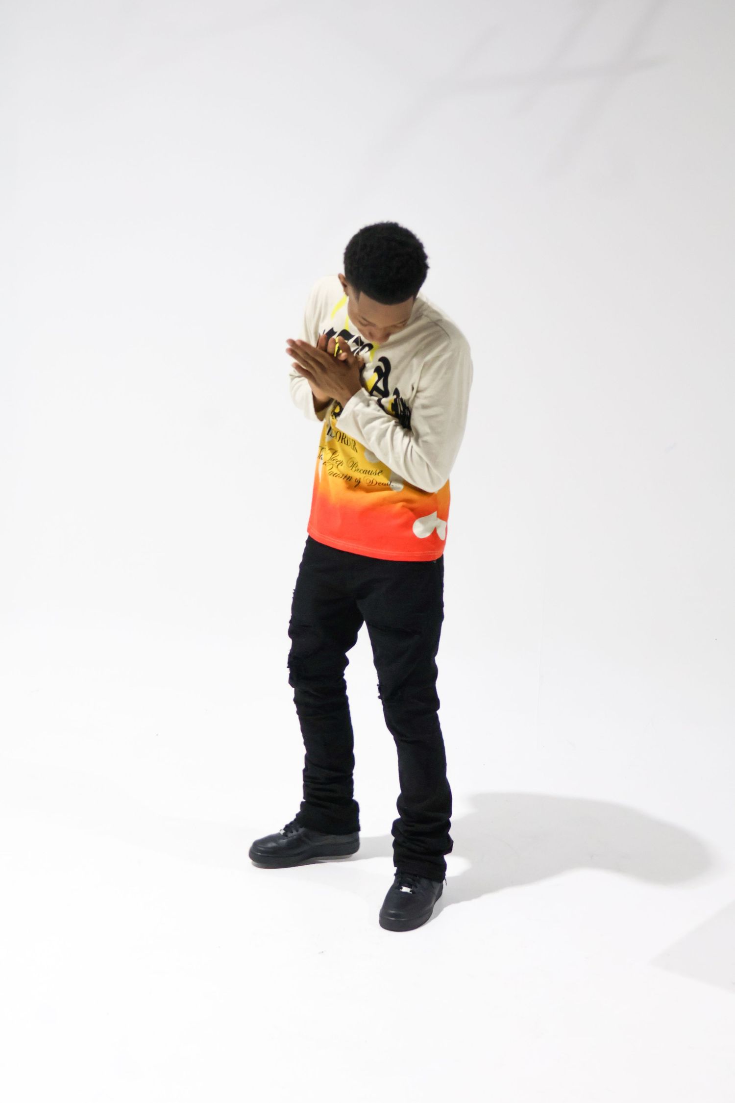 Young man in colorful gradient orange sweatshirt and black jeans poses with hands clasped in white studio.