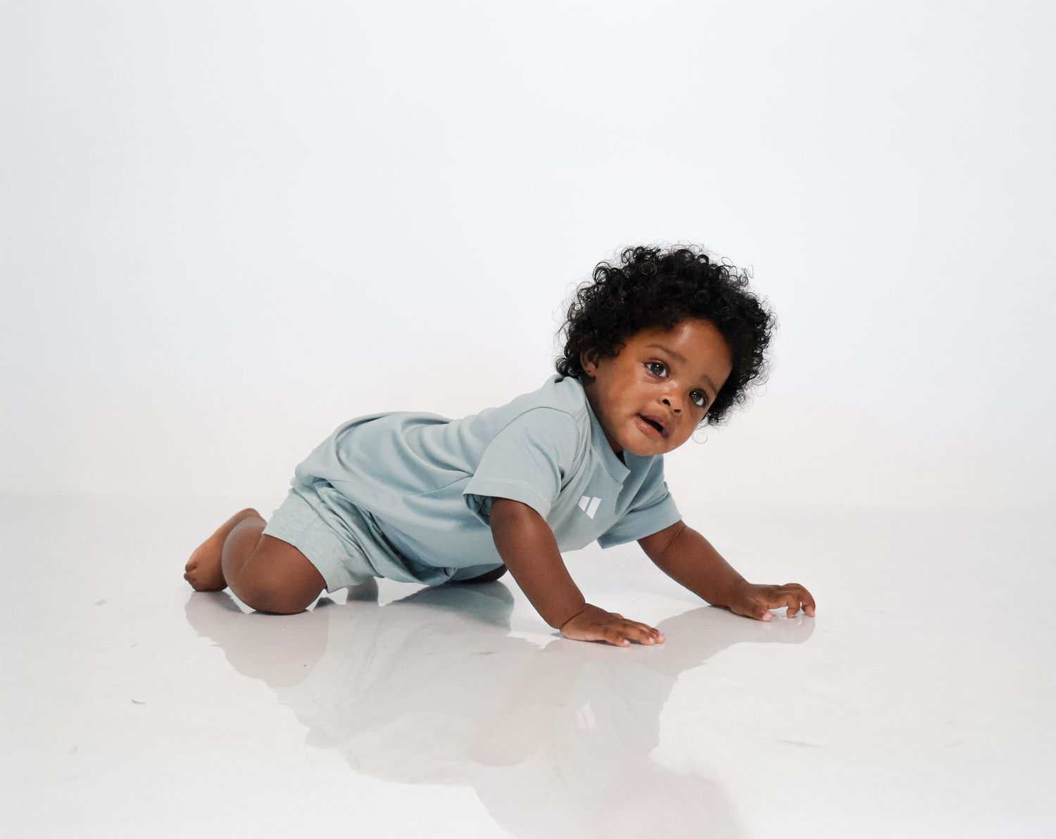 Cute Black baby with curly hair crawling on white floor wearing a light blue romper.
