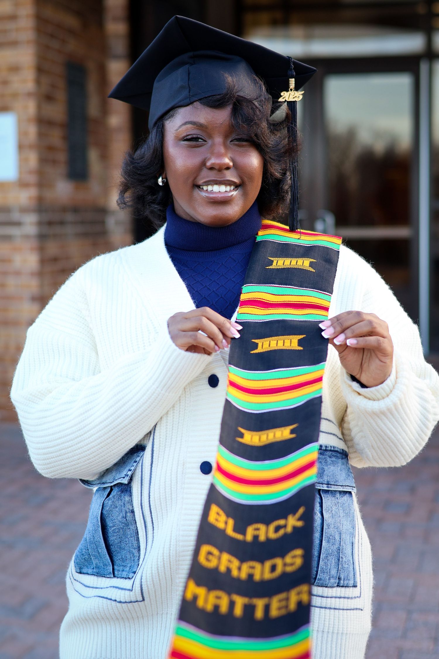 Proud graduate holding Black Grads Matter kente stole outside school building wearing cap and gown.