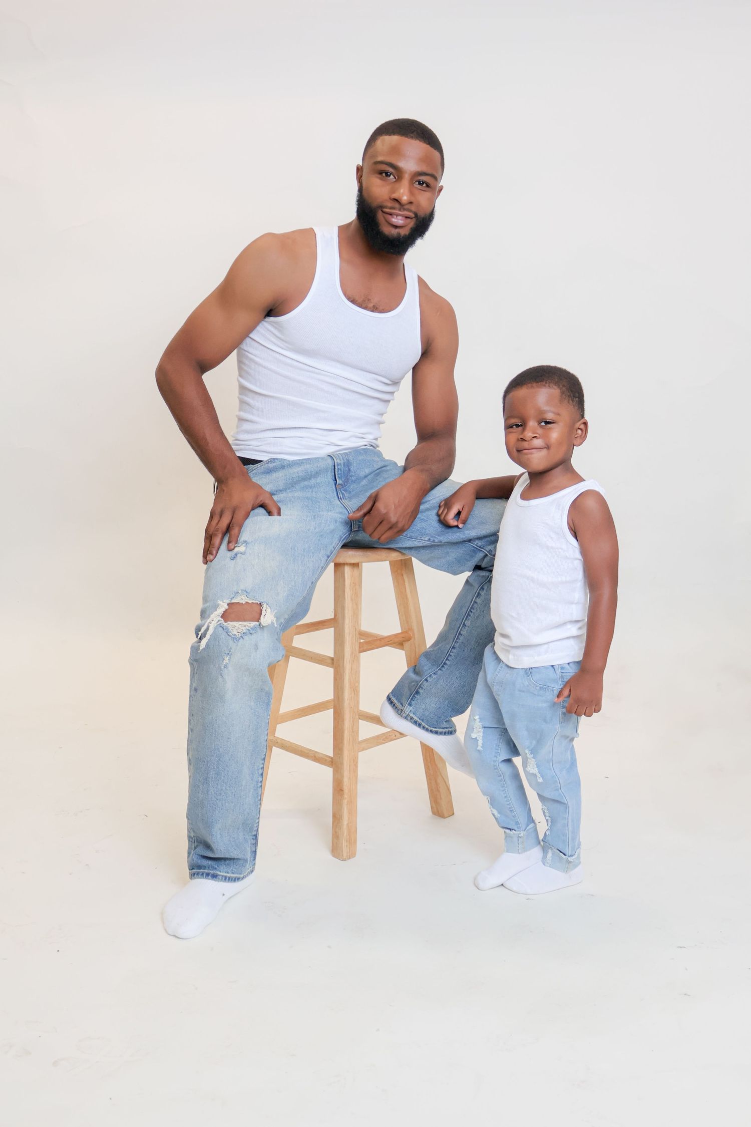 Father and young son in matching white tank tops and ripped jeans posing in a studio portrait.