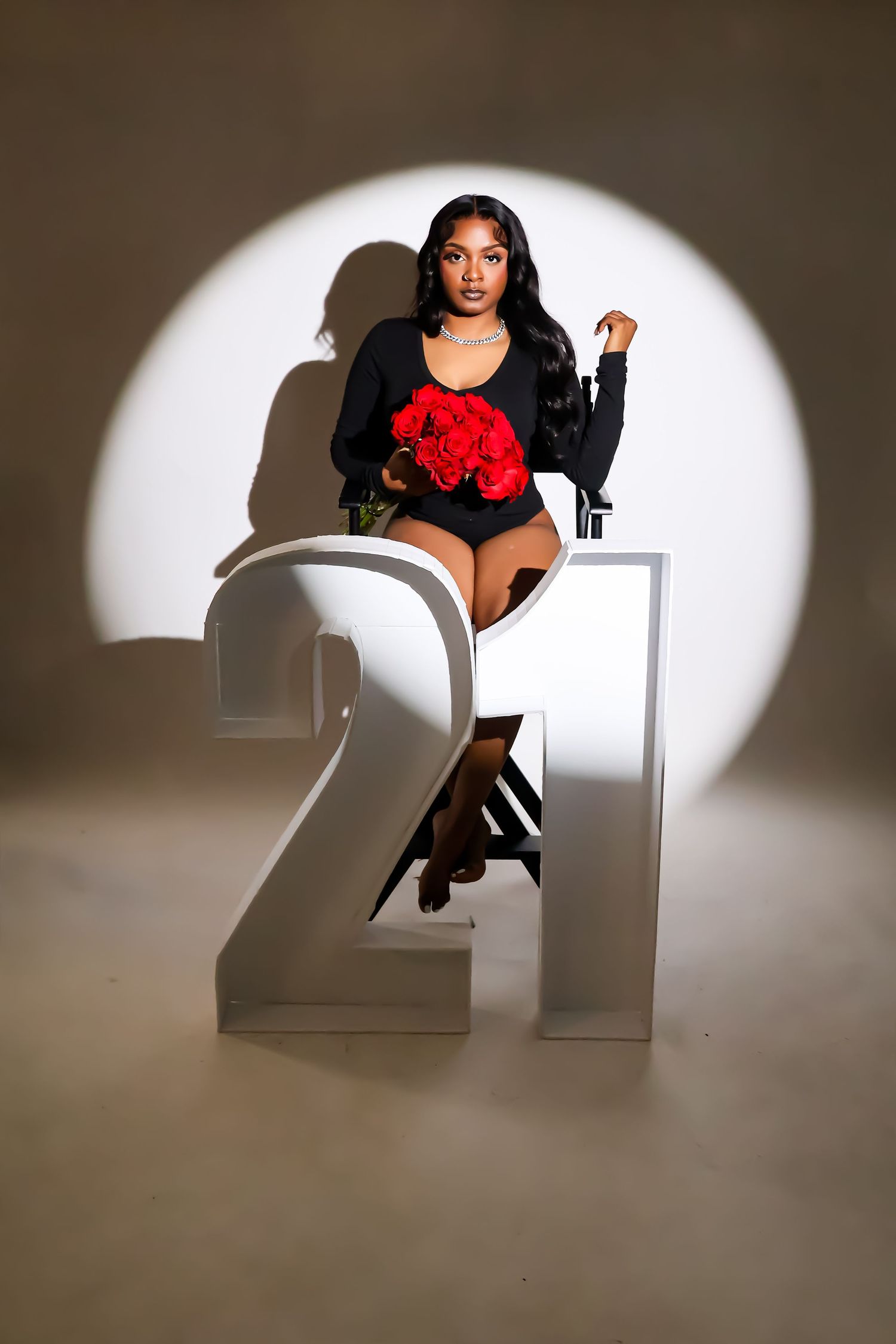 Young woman in black bodysuit holding red roses seated on a chair beside large number 21 prop in spotlight.