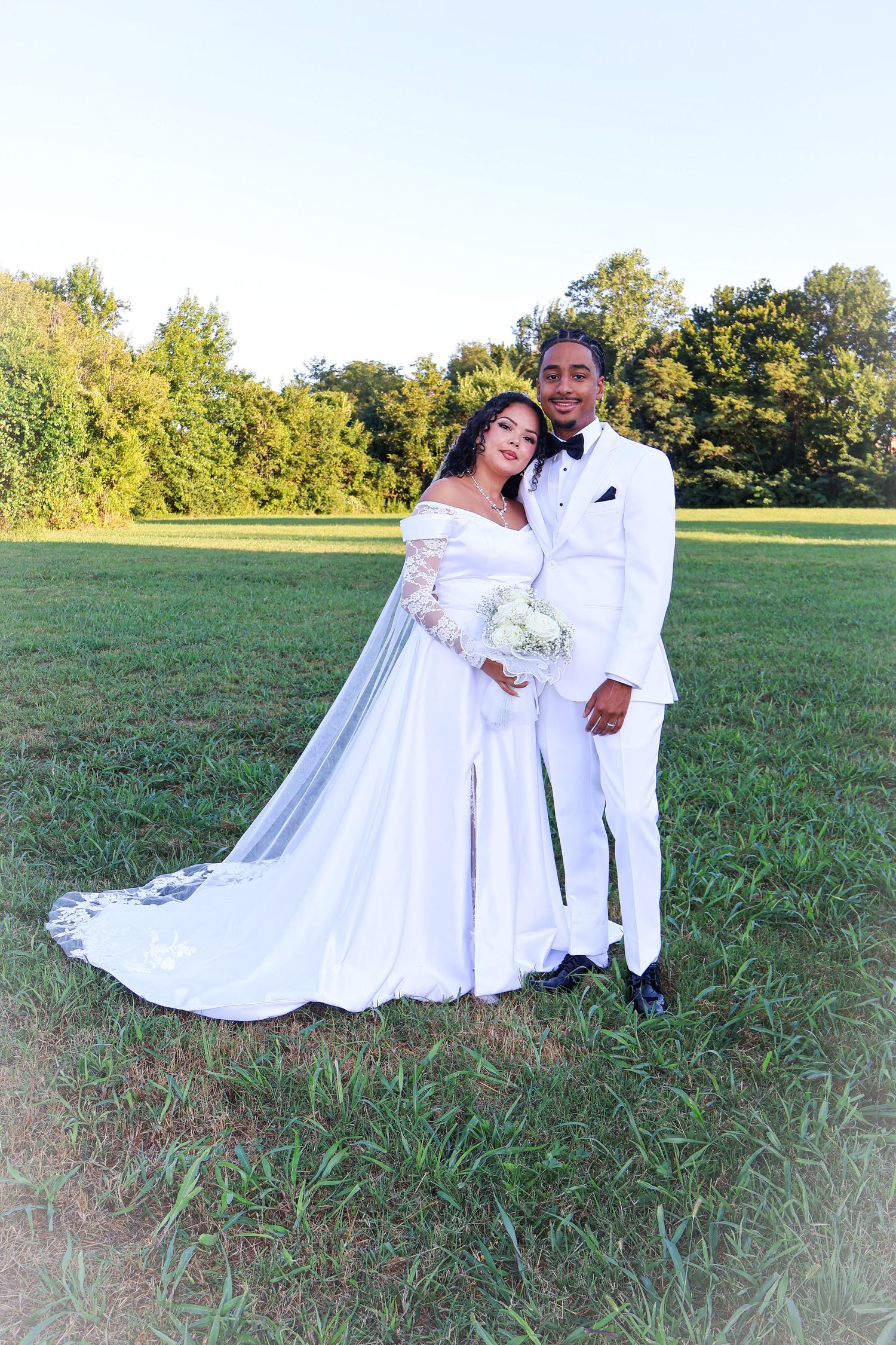 Bride in off-shoulder lace gown with veil and groom in white tuxedo pose in sunlit green field.