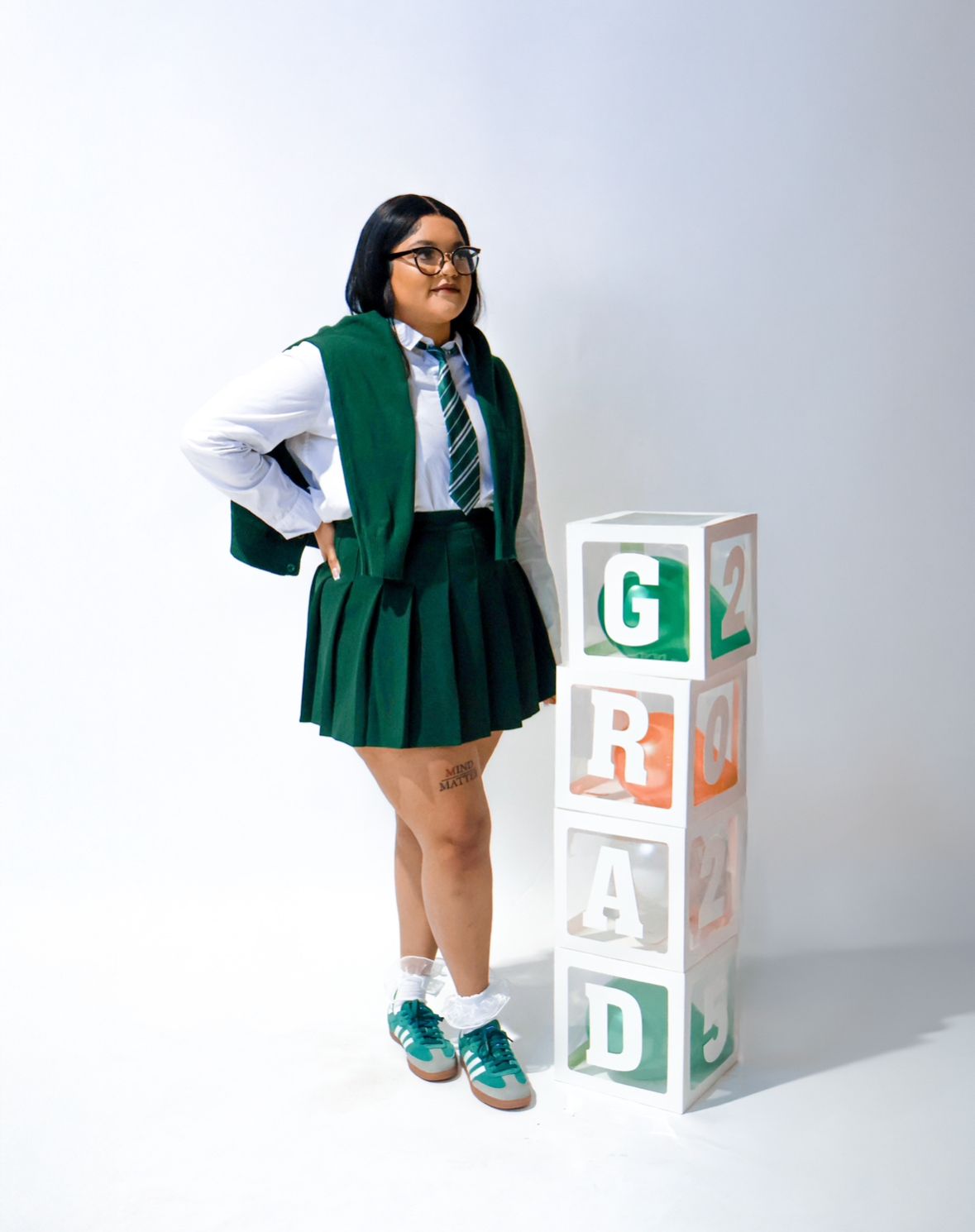 Graduate in green uniform leans on stacked GRAD letter blocks on white background.