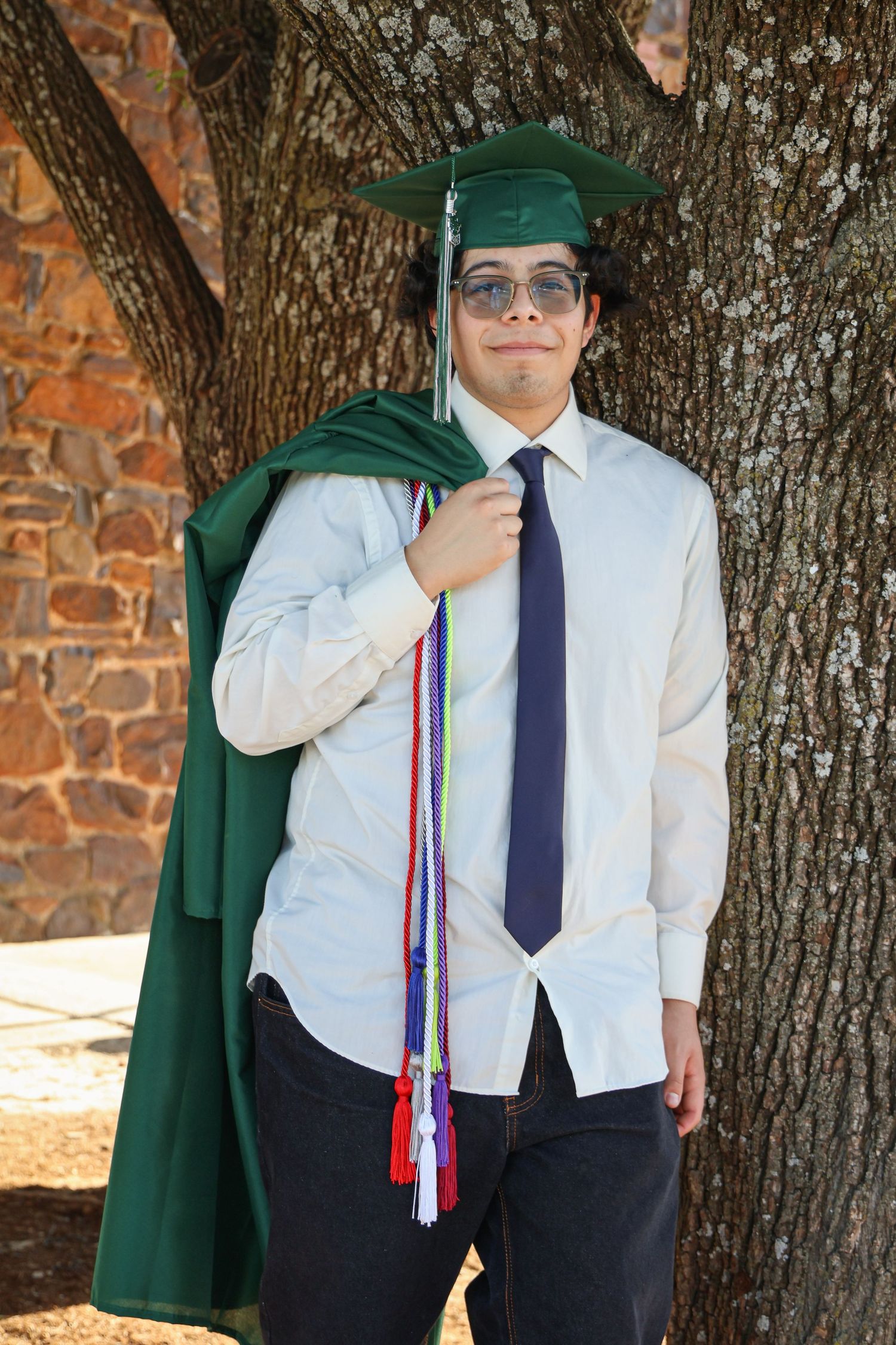 Young man in green graduation cap and gown holding colorful honor cords, posing by a tree in white shirt and tie.