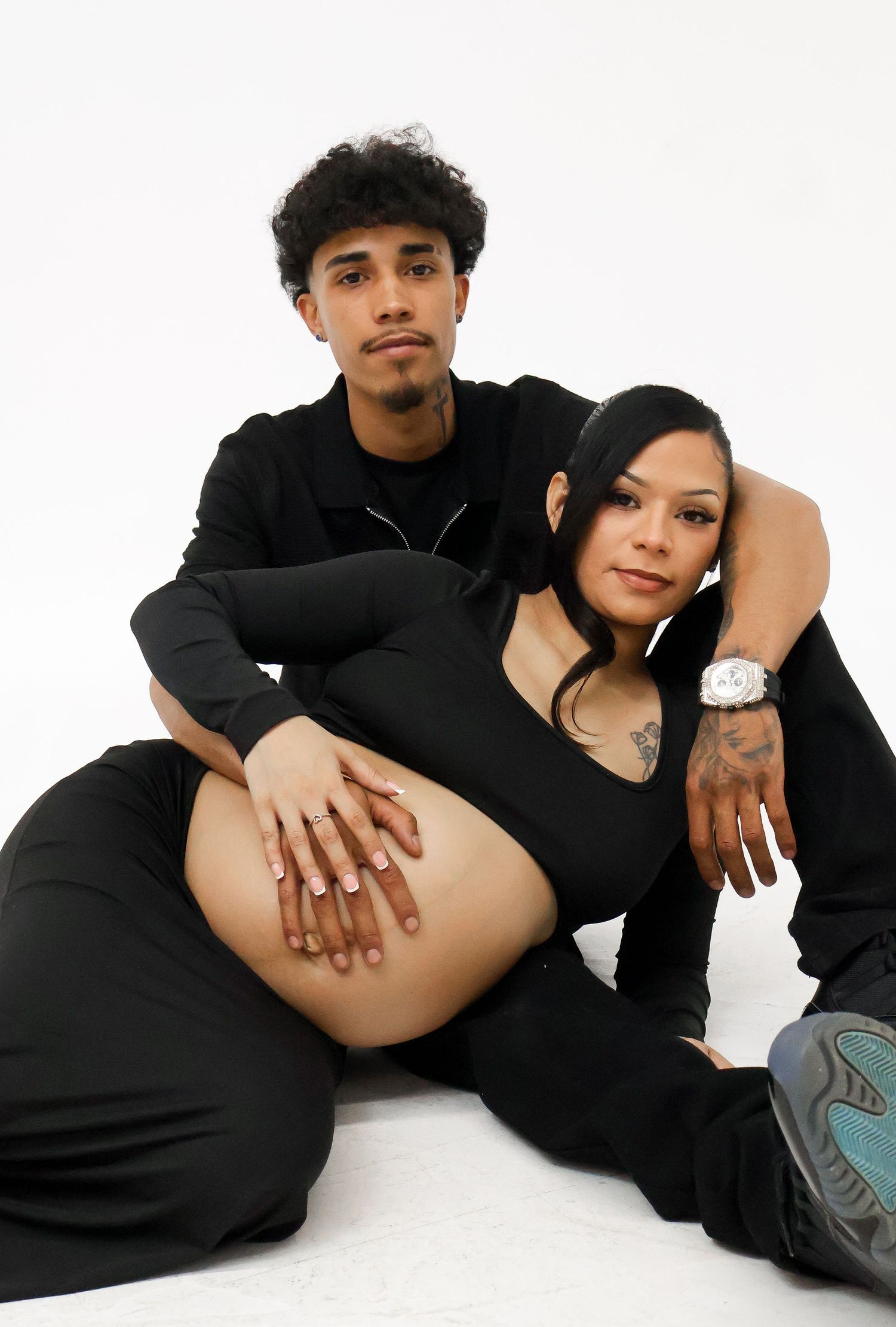 Couple in black outfits pose together on floor showcasing pregnant belly in maternity shoot.