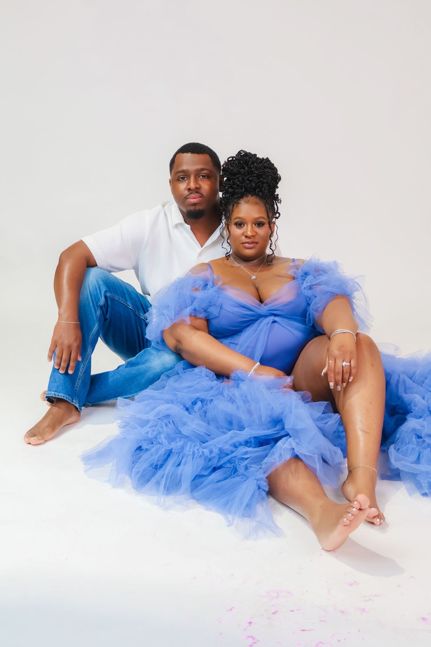 Couple posing for maternity photo, woman in blue tulle gown, man in white shirt and jeans, white studio background.
