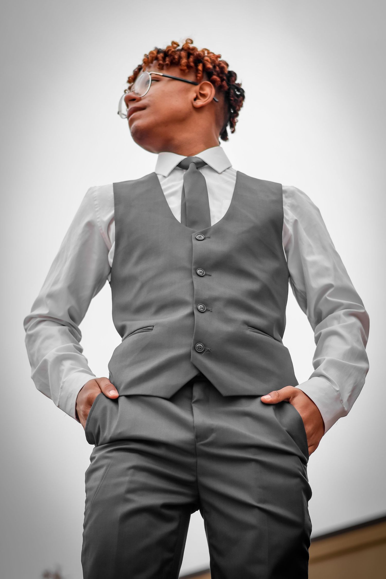 Confident young Black man in gray vest and tie poses with hands in pockets outdoors.