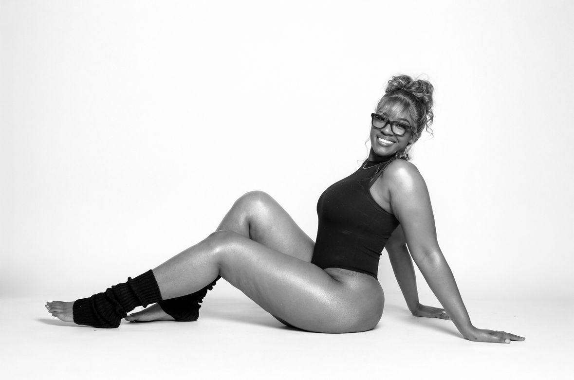 Woman in black bodysuit sitting on floor posing in black and white studio portrait.