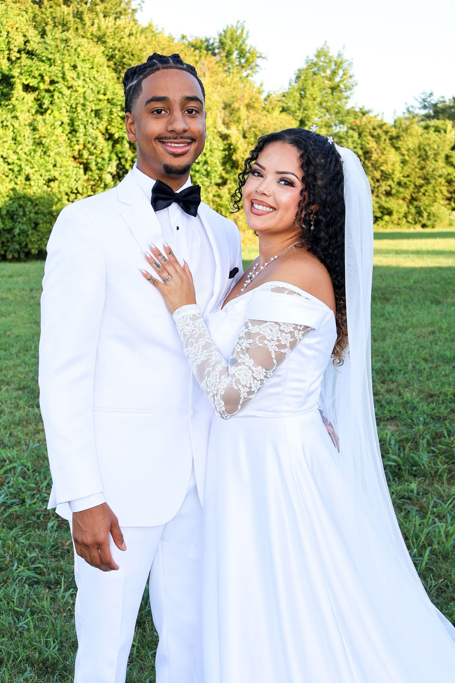 Happy newlywed couple posing outdoors, groom in white tuxedo and bride in elegant off-shoulder wedding gown with veil.