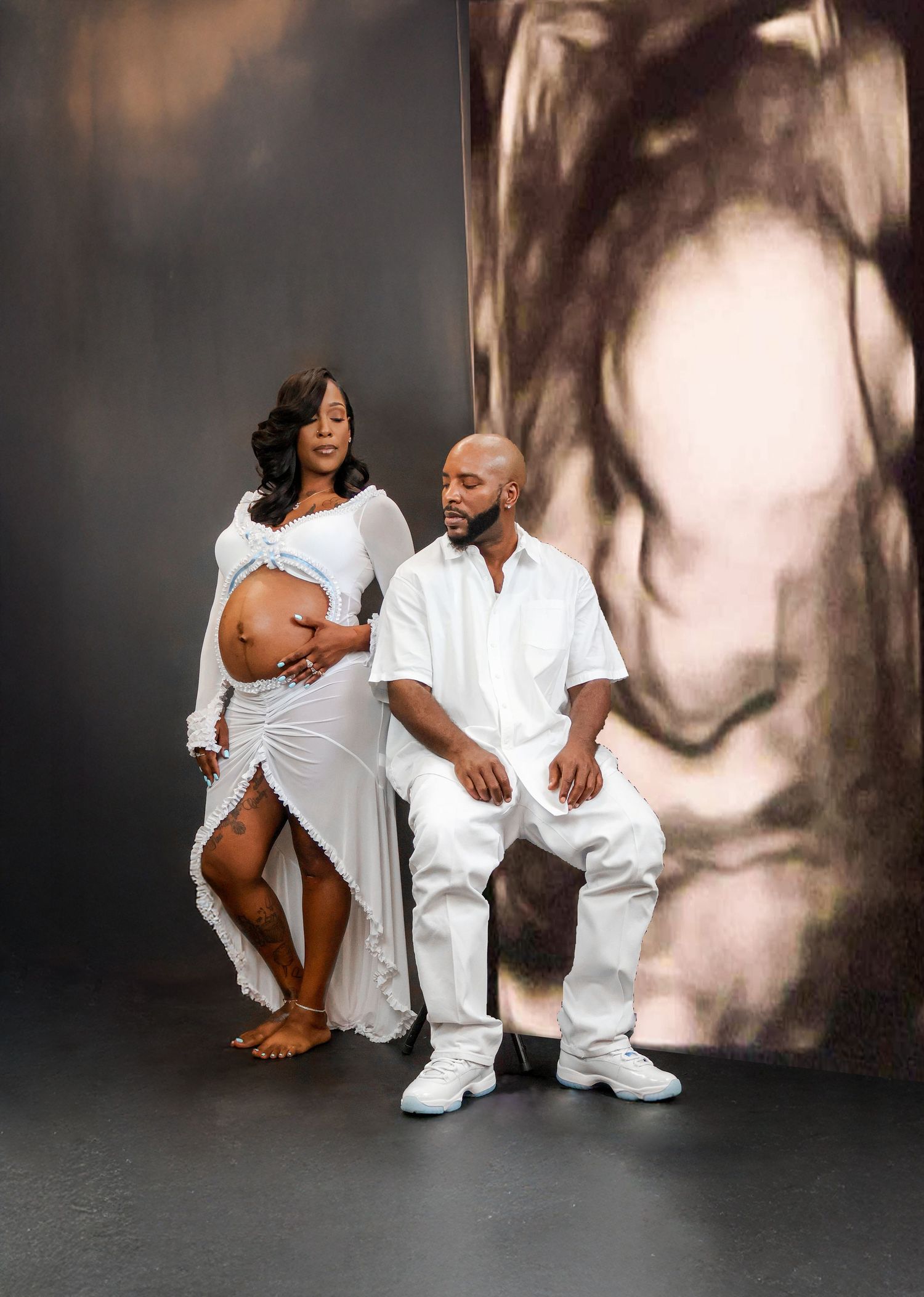 Couple in white outfits poses for maternity photoshoot with ultrasound backdrop, woman showing baby bump.