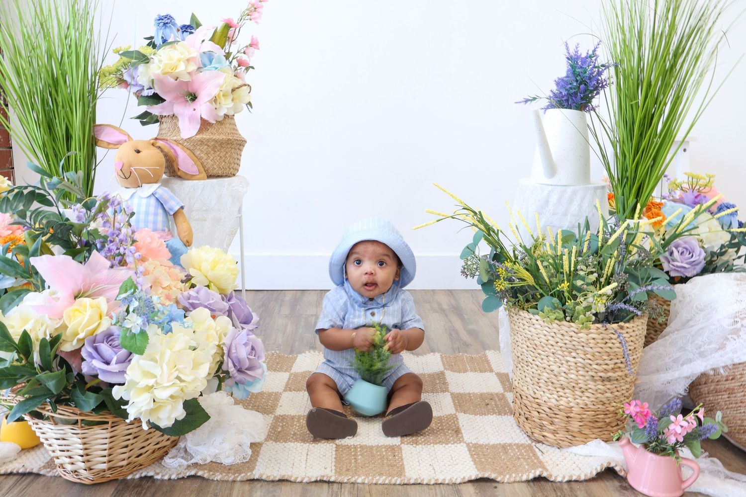 Cute baby in blue hoodie sitting among colorful spring flower arrangements in Easter photo session.