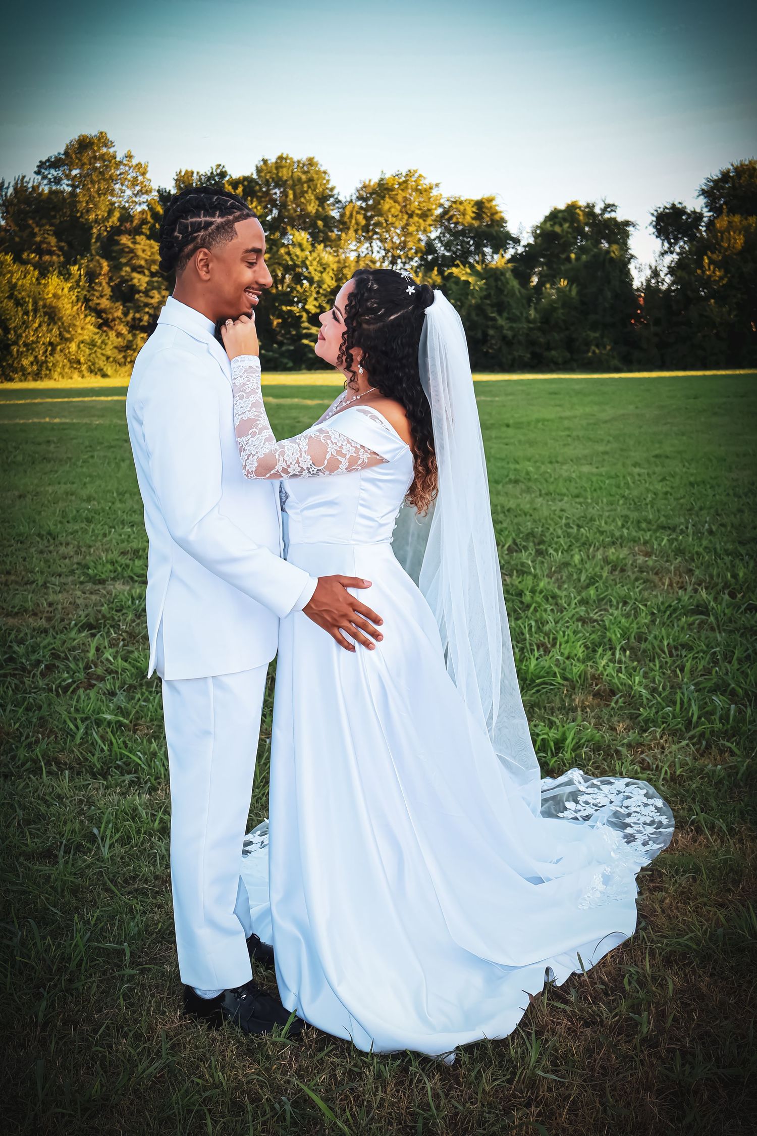 A bride and groom in white attire share a romantic moment in a lush green field with trees in the background.