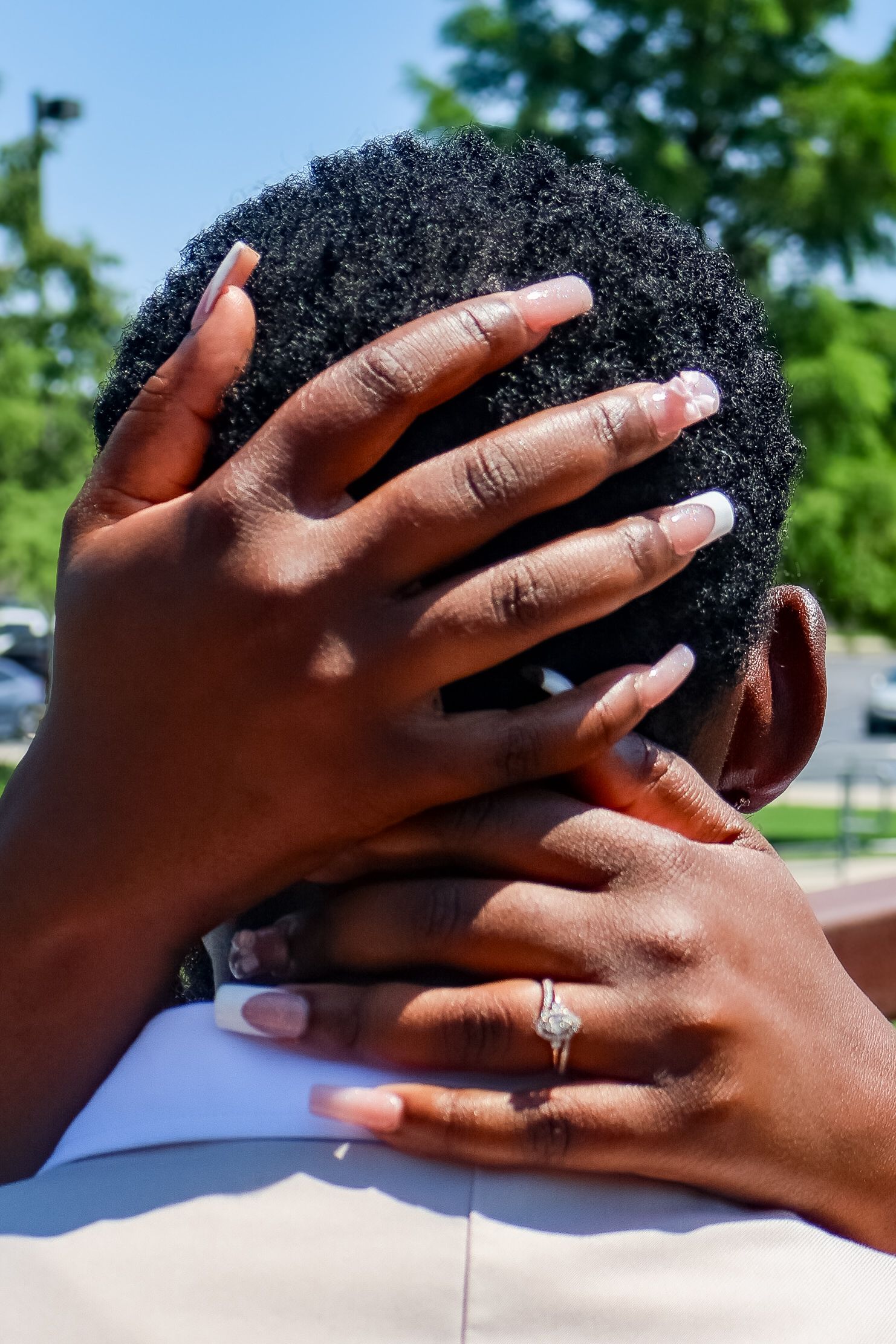Close-up of hands with manicured nails and diamond engagement ring embracing mans head.