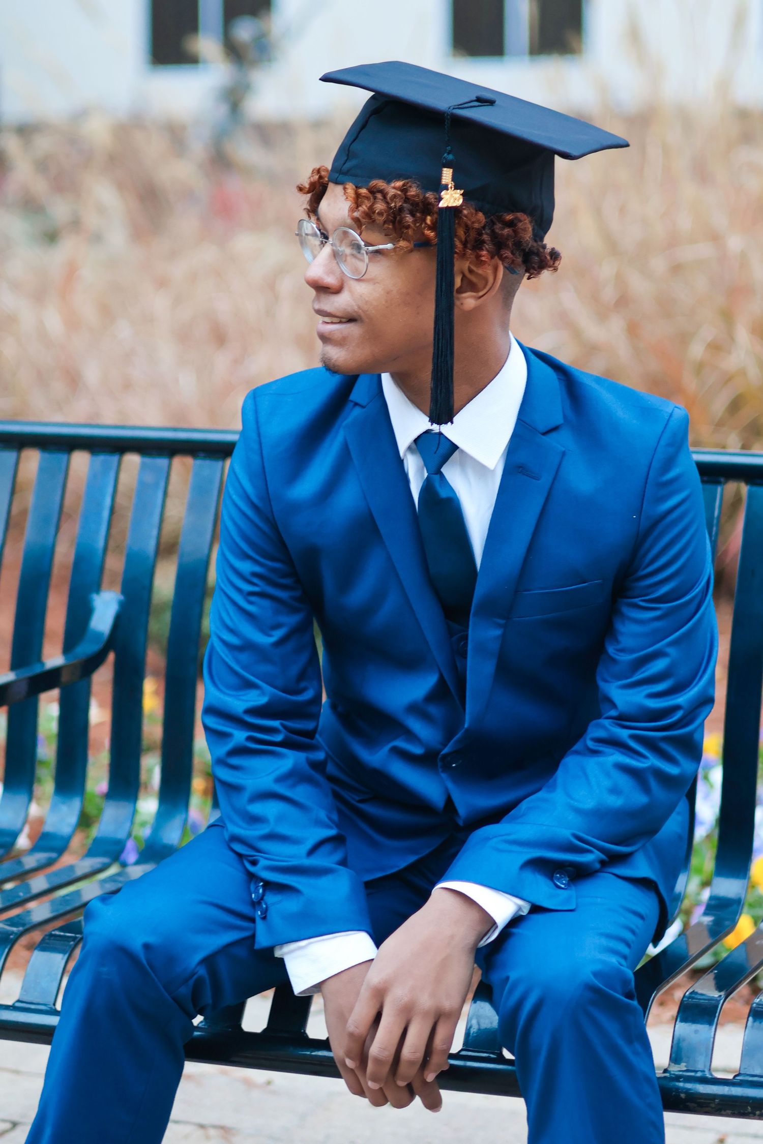 Young Black male graduate in blue suit and cap sits on bench looking away outdoors.