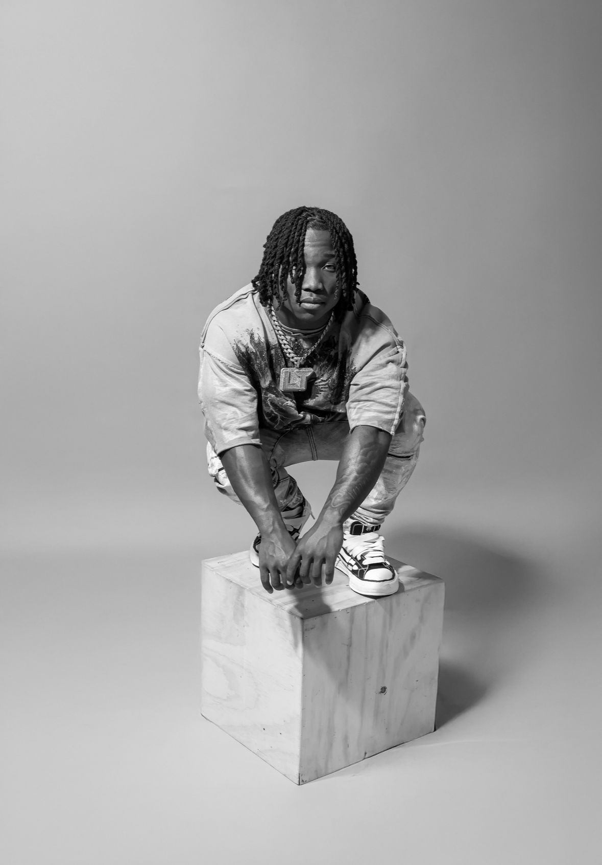 Black and white photo of a young man with braids crouching on a wooden cube, wearing chains and sneakers.