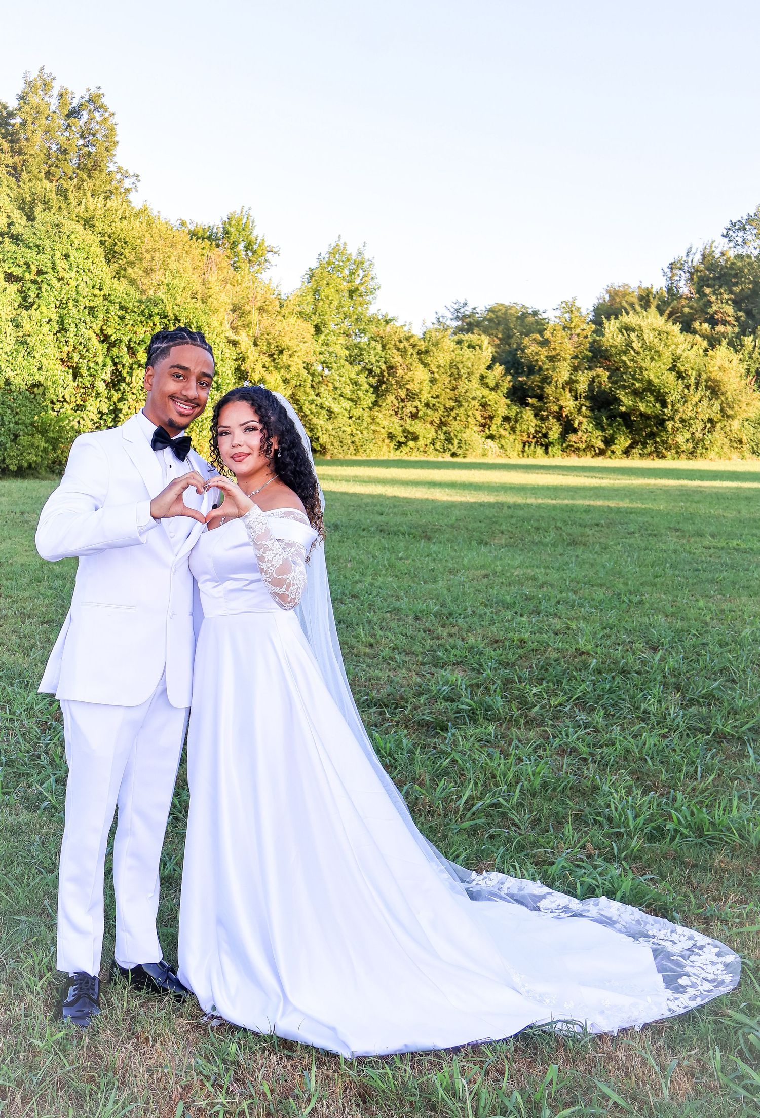 Newlywed couple in matching white wedding attire making a heart shape with their hands outdoors on a green lawn.