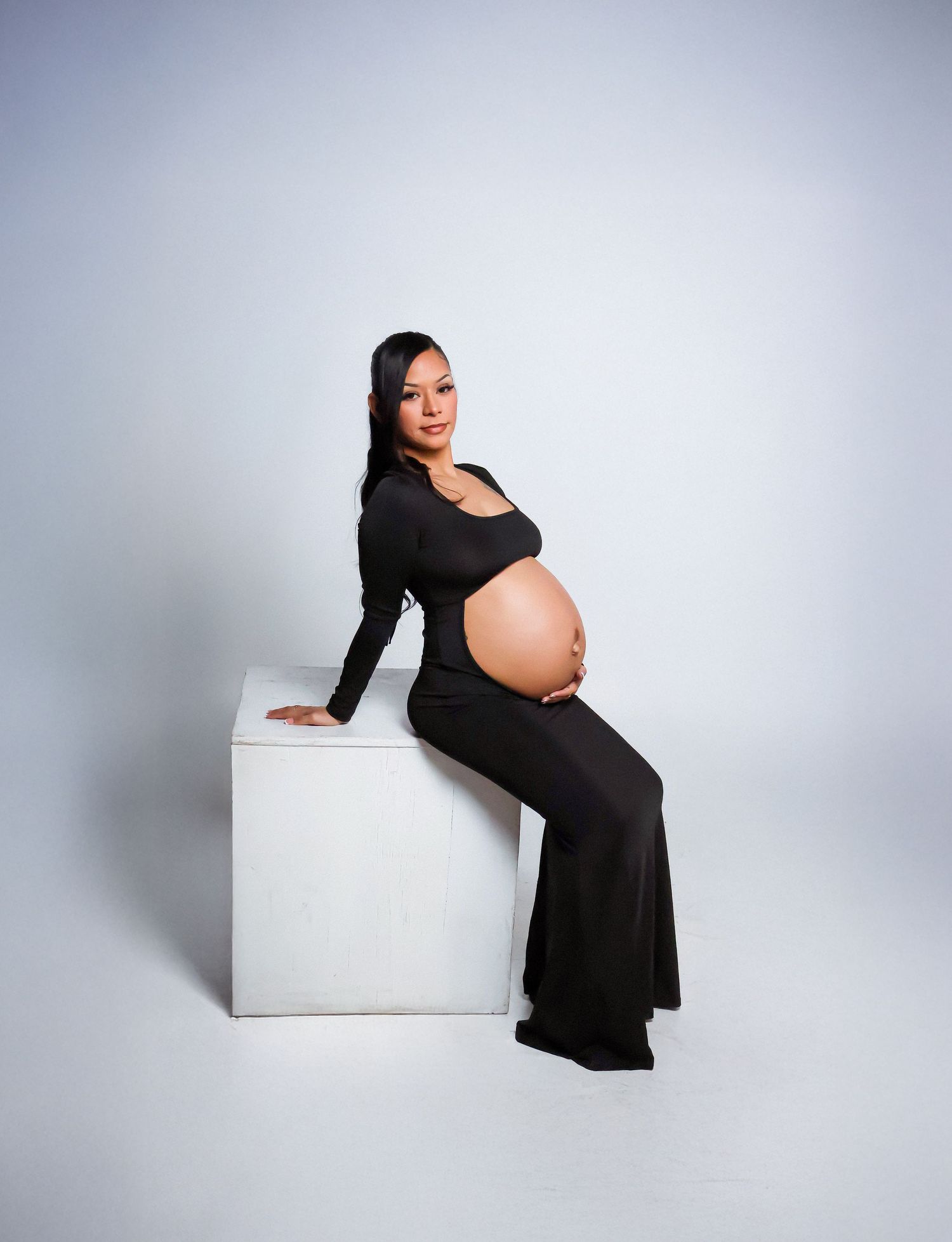 Pregnant woman in black maternity gown sitting on white cube, cradling bare baby bump in professional studio photo.