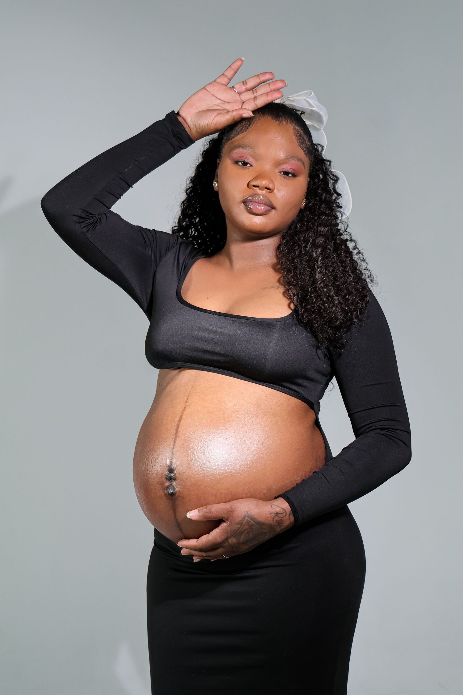 Pregnant Black woman in black crop top and skirt cradling baby bump, posing gracefully in studio setting.