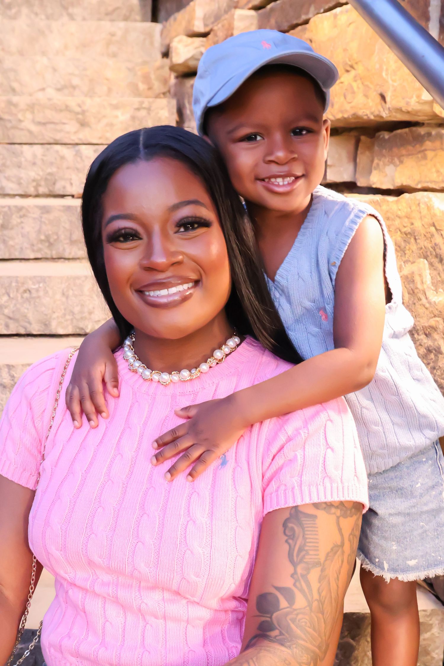 A smiling woman in a pink top with a pearl necklace poses with a young child in a blue cap on outdoor steps.