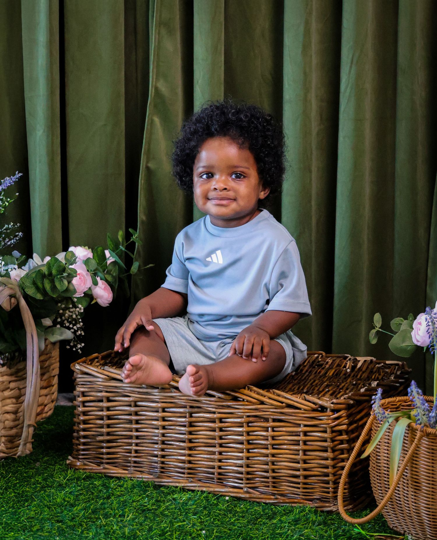 Adorable baby boy in blue Adidas outfit sitting in a wicker basket with flowers on green grass.