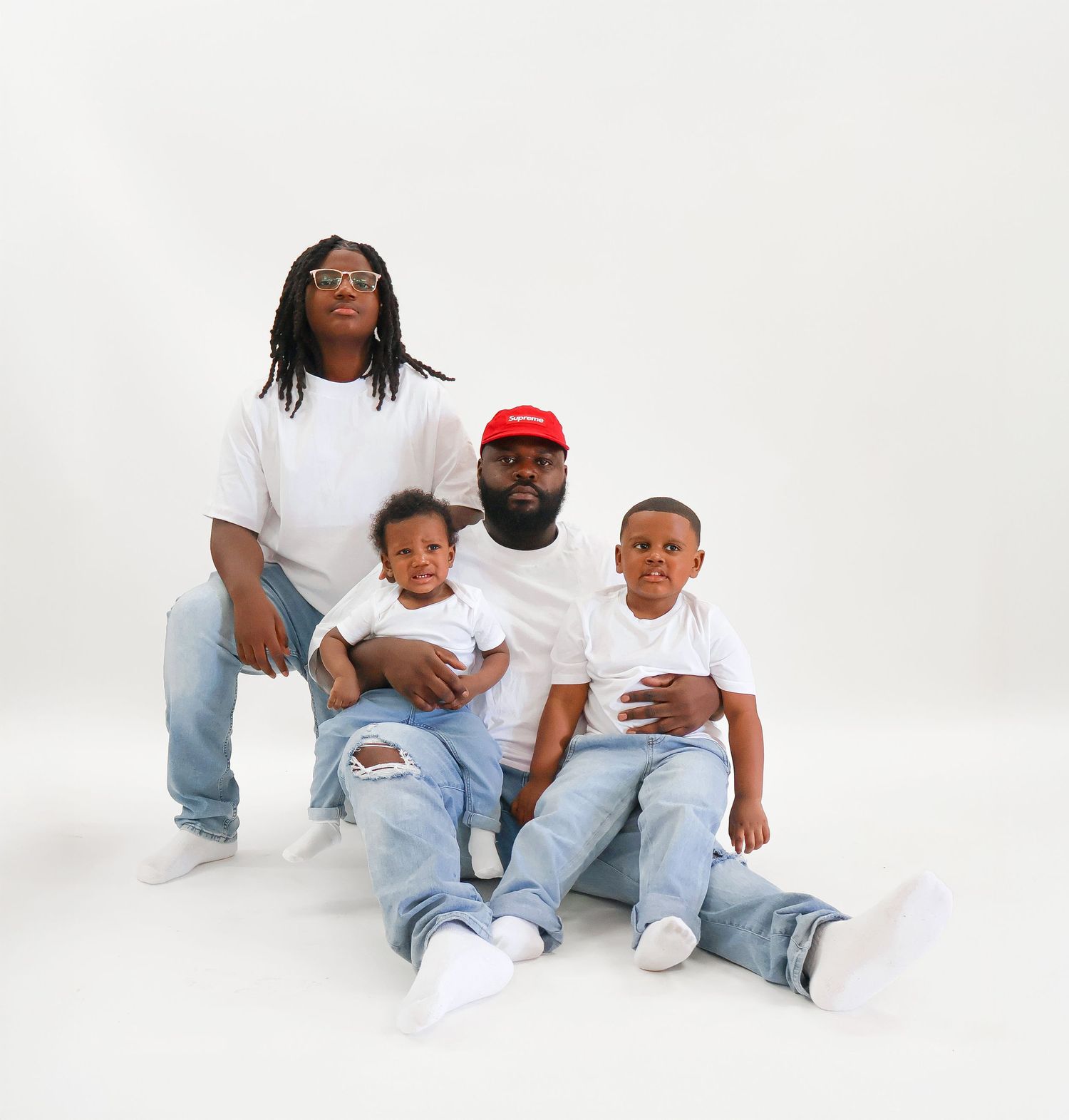 A father and three sons dressed in white shirts and jeans pose together in a clean studio portrait.