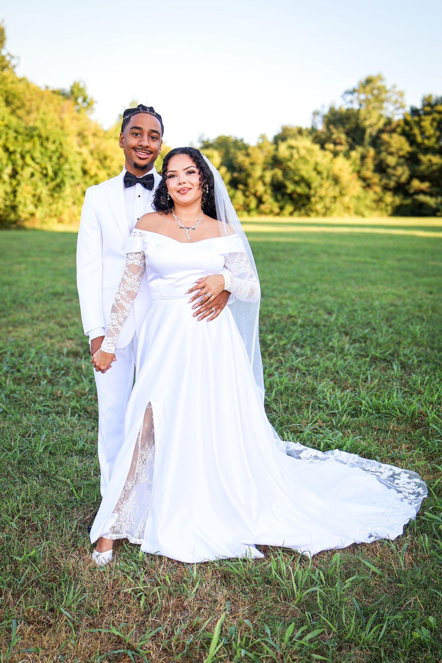 Smiling bride and groom in white wedding attire pose together on lush green lawn at sunset.