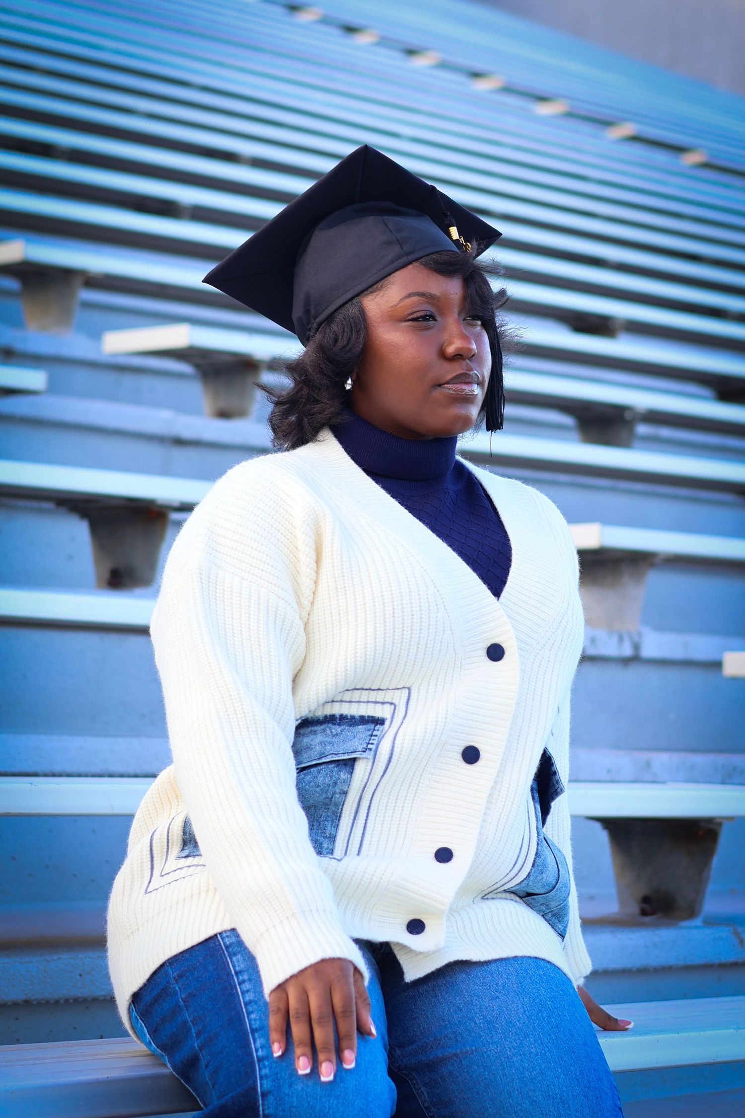 Graduate in mortarboard cap and white cardigan sitting thoughtfully on stadium bleachers.