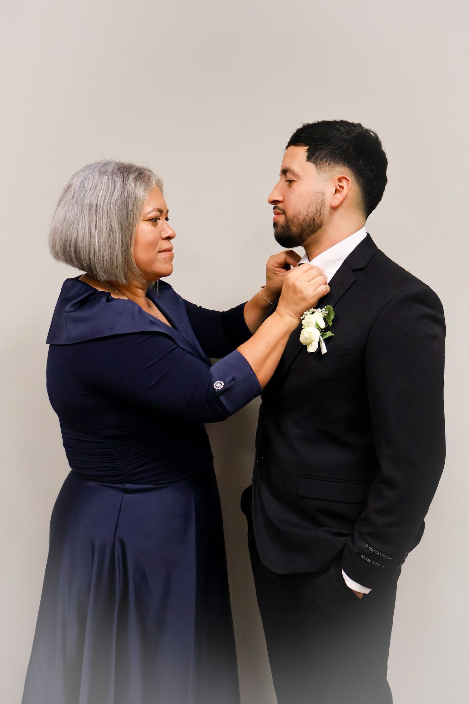 Older woman in navy dress adjusts the tie of a groom in a black suit with boutonniere.
