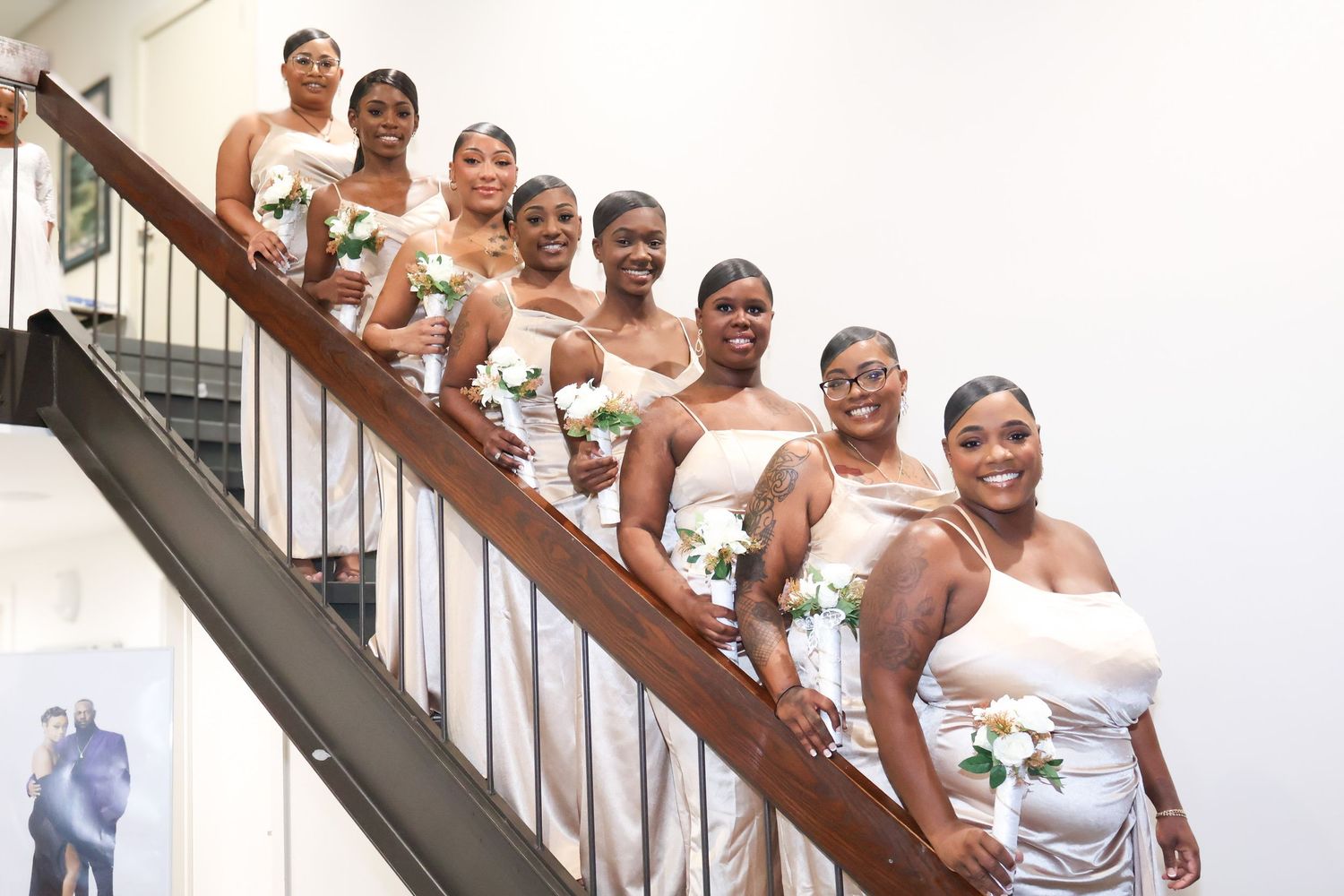 Eight bridesmaids in silver gowns holding white roses pose on a staircase for wedding portrait.