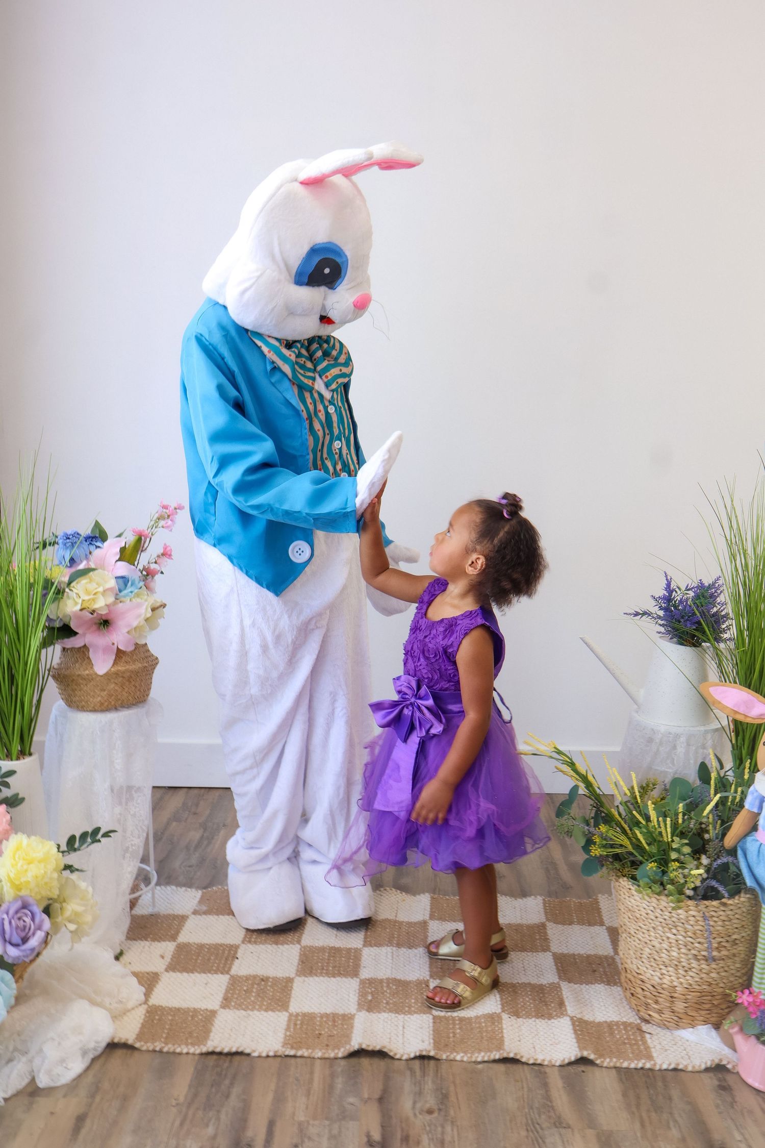 Young girl in purple dress high-fiving Easter Bunny mascot in festive spring decorated photo studio.