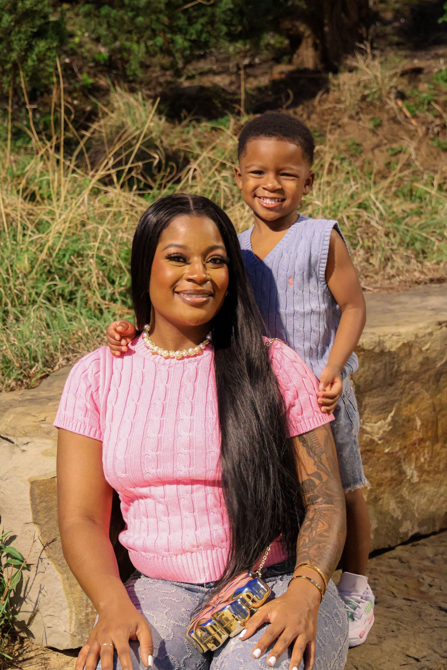A smiling woman in a pink top sits outdoors with a young boy standing behind her, both posing happily.