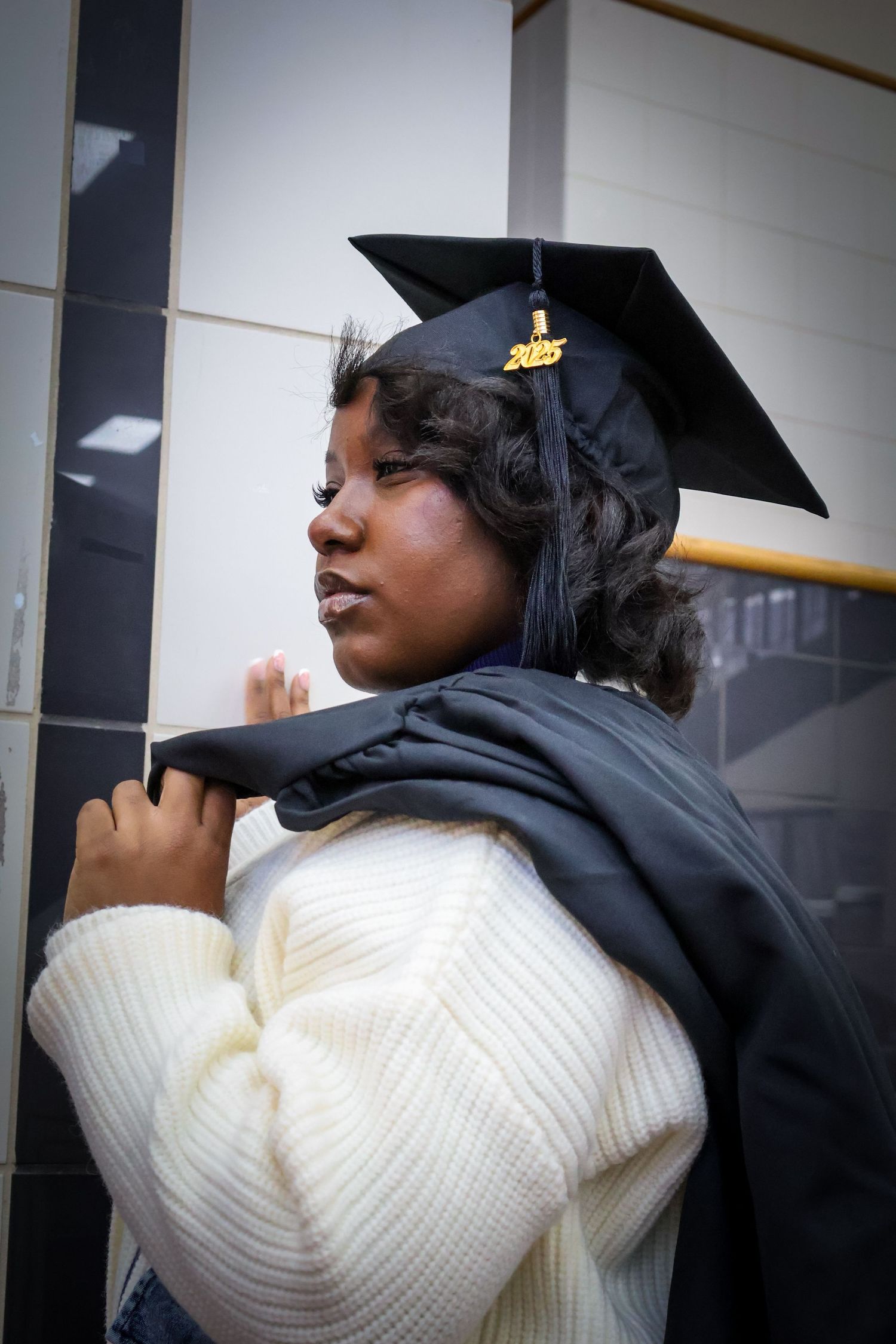Young Black woman in graduation cap and gown over white sweater poses thoughtfully indoors.