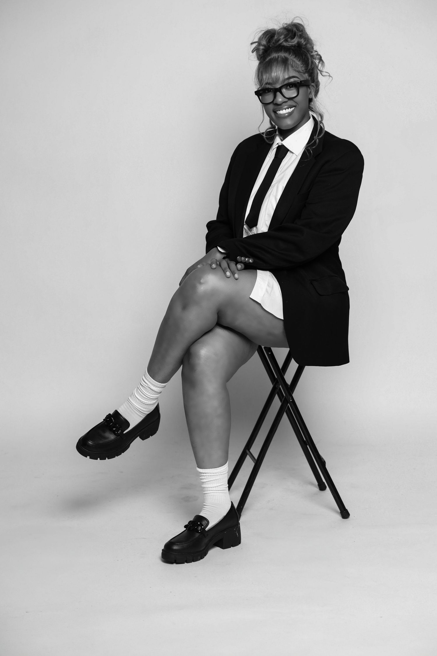 Woman in black blazer and tie sitting on stool smiling in black and white photo.