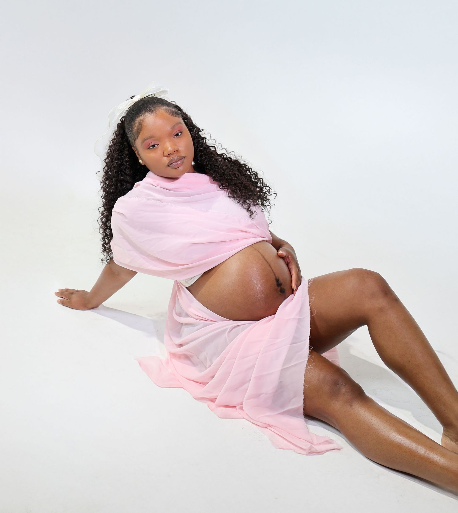 Pregnant woman in pink sheer wrap fabric posing on white background for maternity photoshoot.