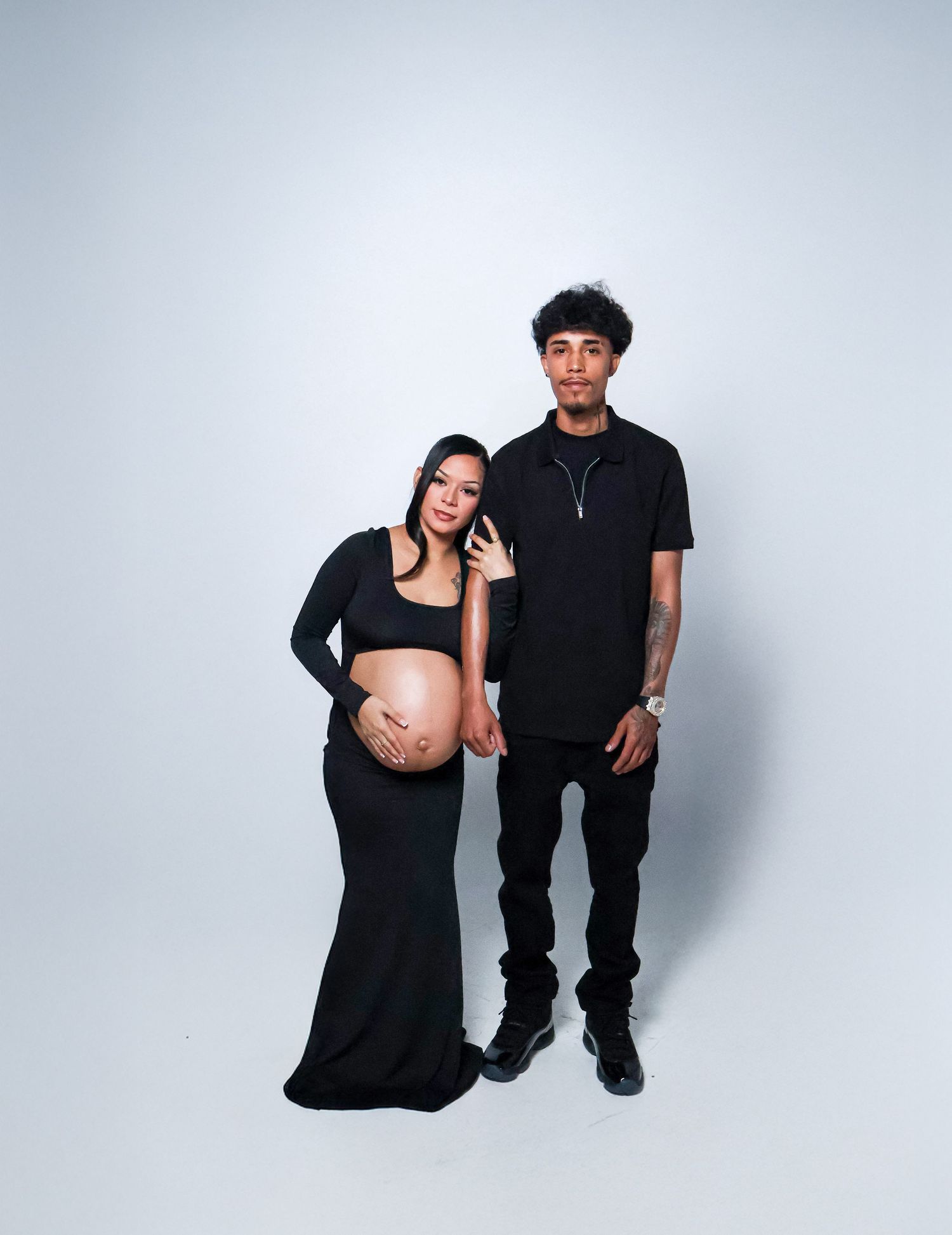Pregnant couple in matching black outfits posing for a maternity photo shoot against a white background.