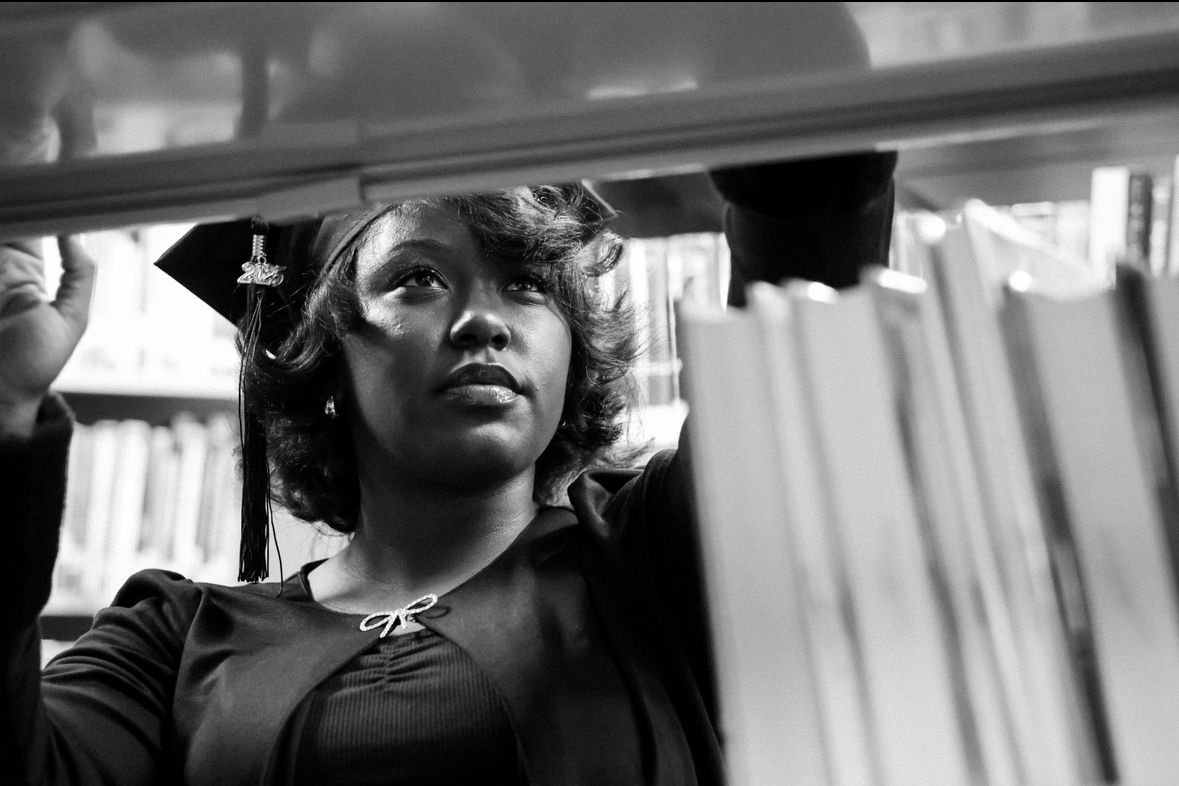 Black and white artistic portrait of graduate in cap browsing books on a library shelf.