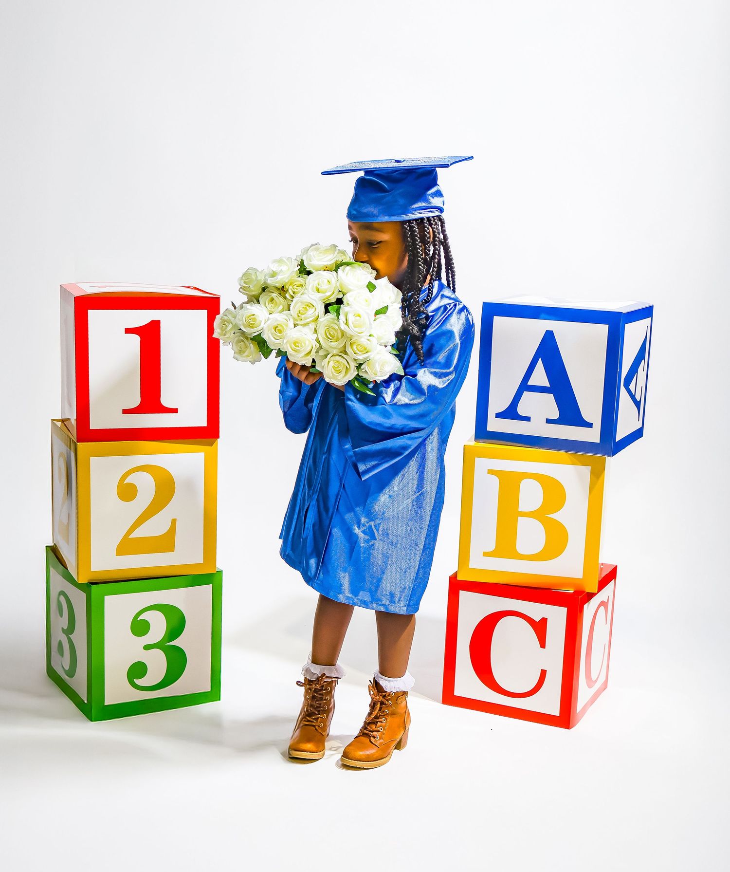 Young child in blue graduation cap and gown holds white flowers beside large colorful ABC and 123 alphabet blocks.