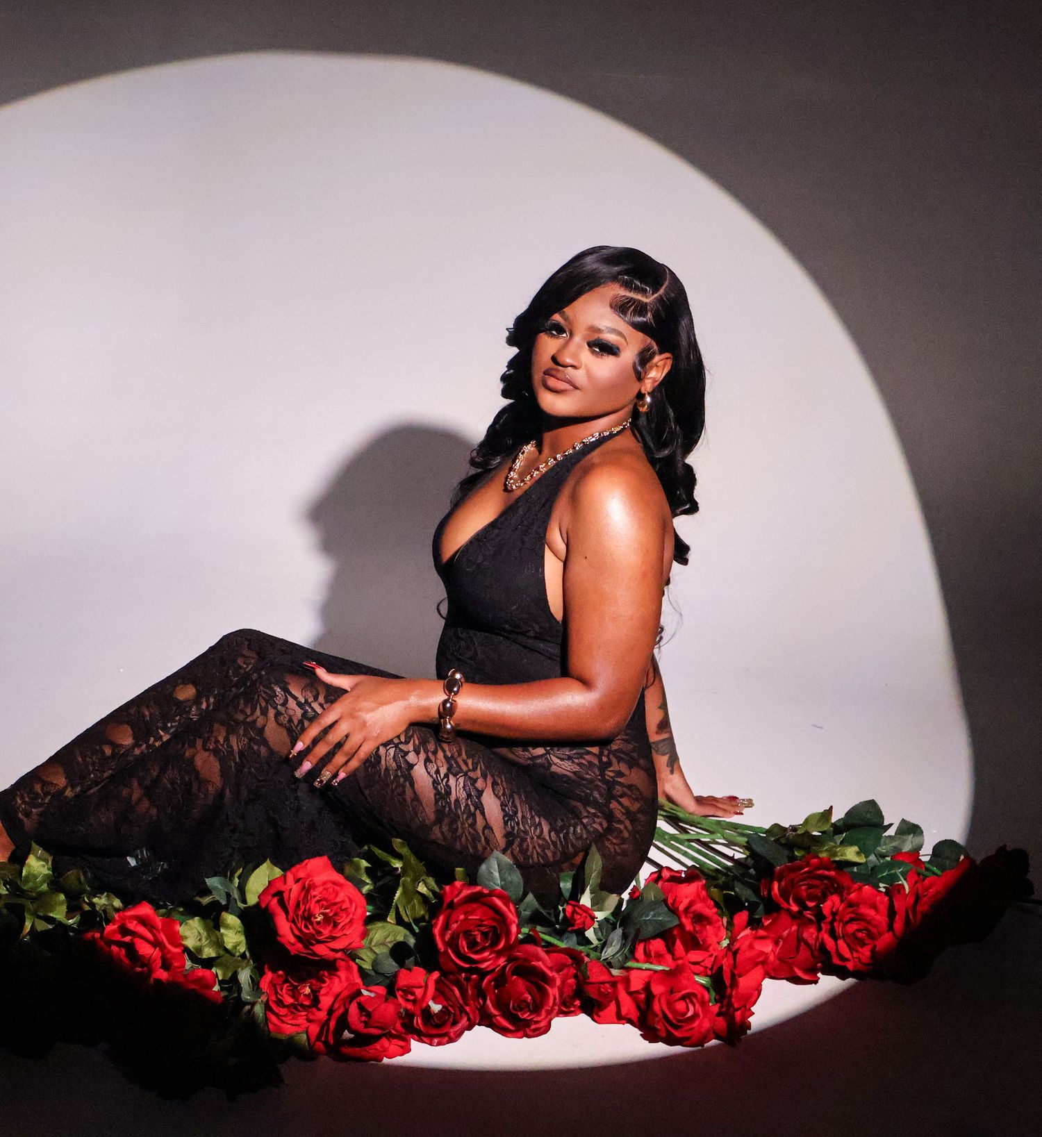 Woman in black outfit posing with red roses on floor in front of a circular light backdrop.