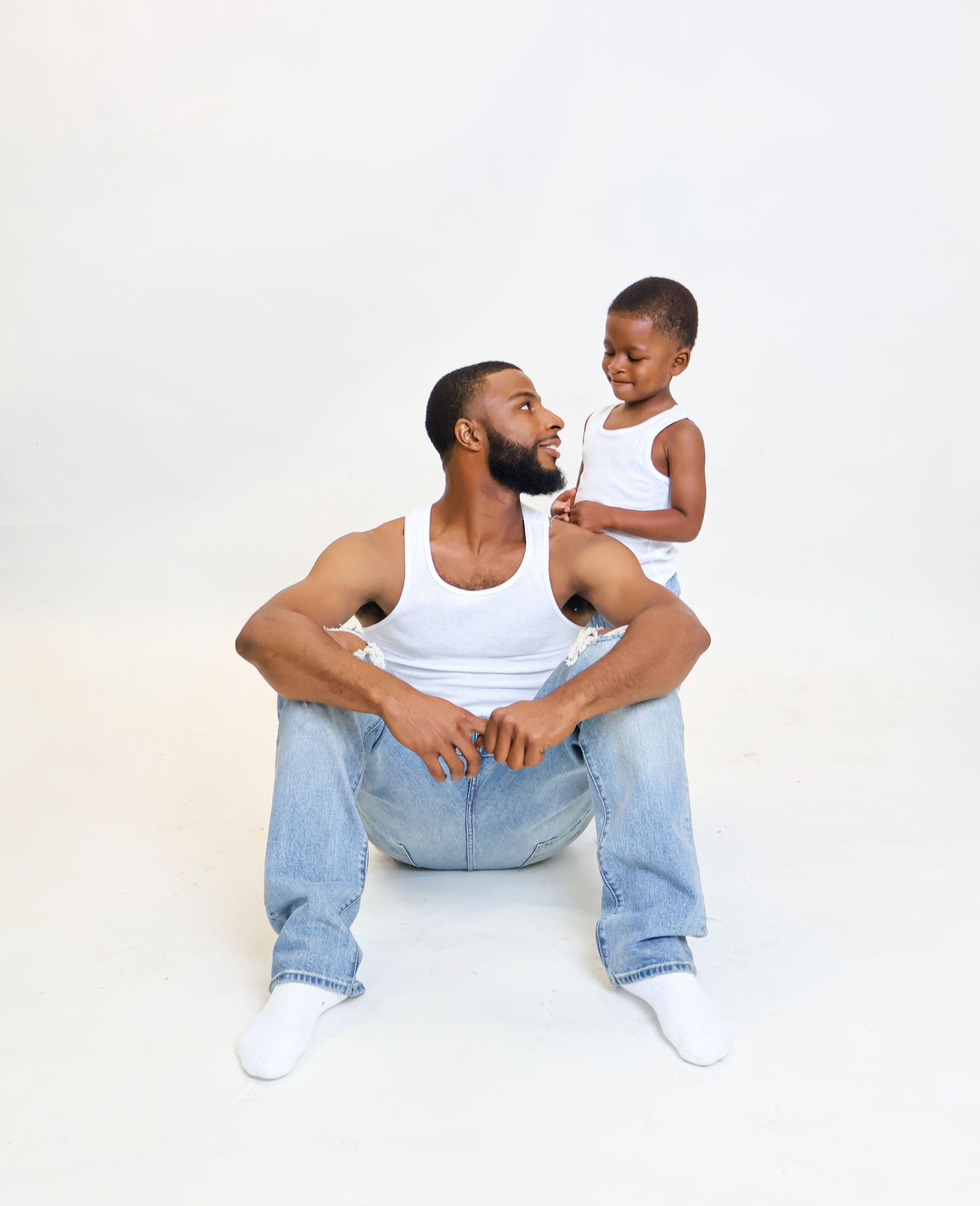 Father sitting on floor while toddler son stands behind him smiling in a white studio setting.