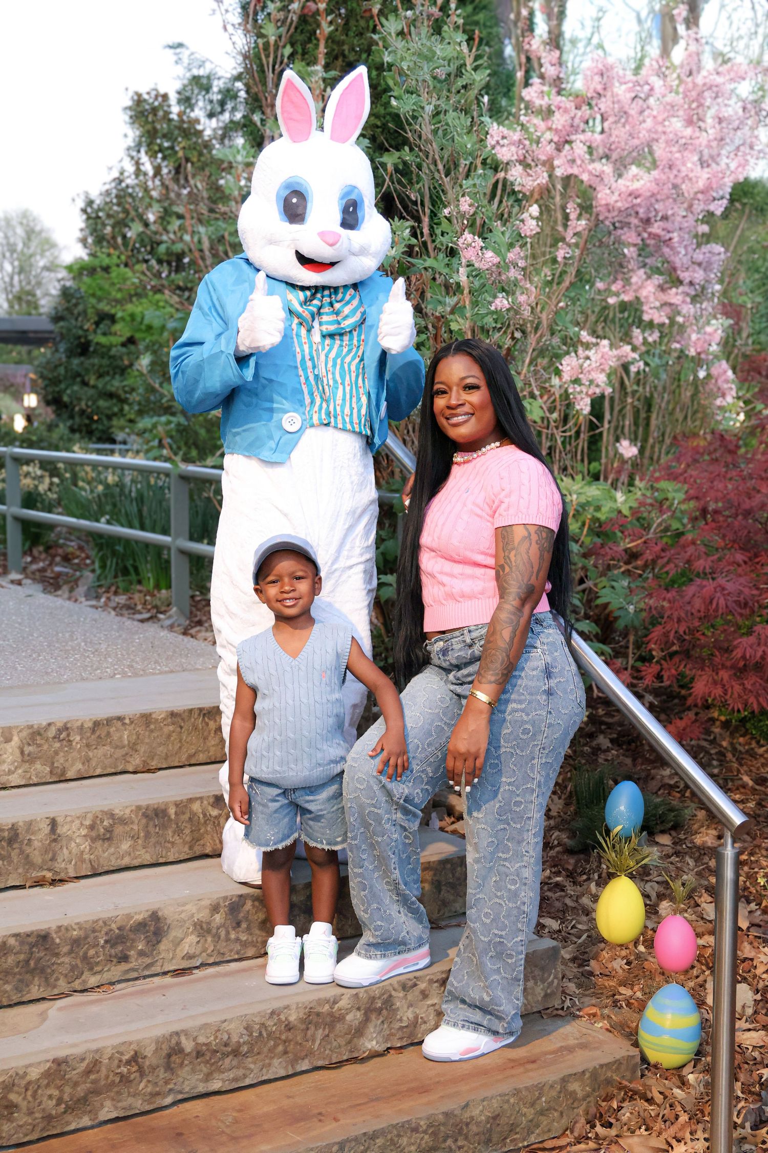 Woman and child pose with Easter Bunny mascot on steps surrounded by colorful Easter eggs and spring flowers.
