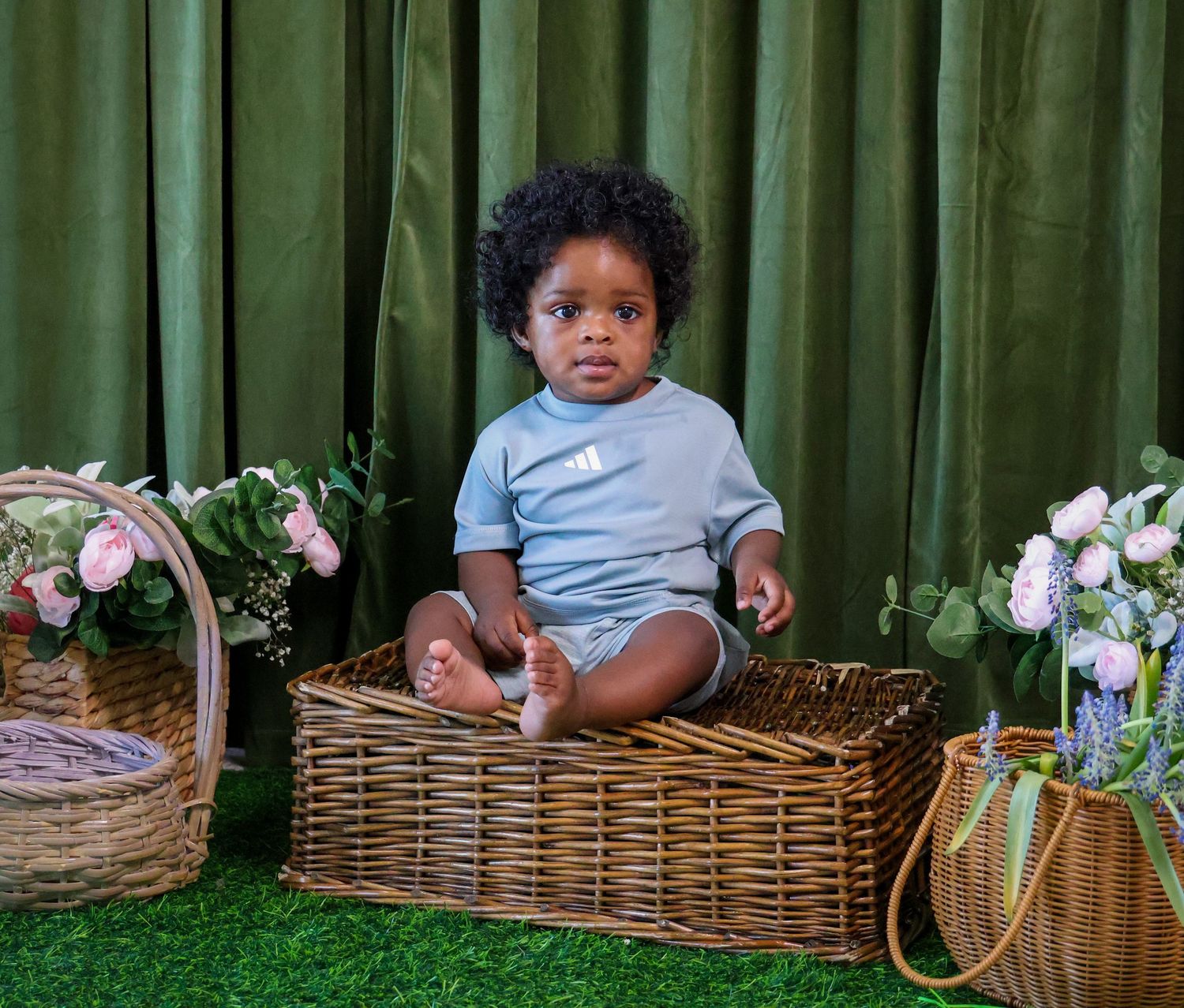 Cute baby in light blue onesie sitting in wicker basket surrounded by flowers and baskets on green grass backdrop.