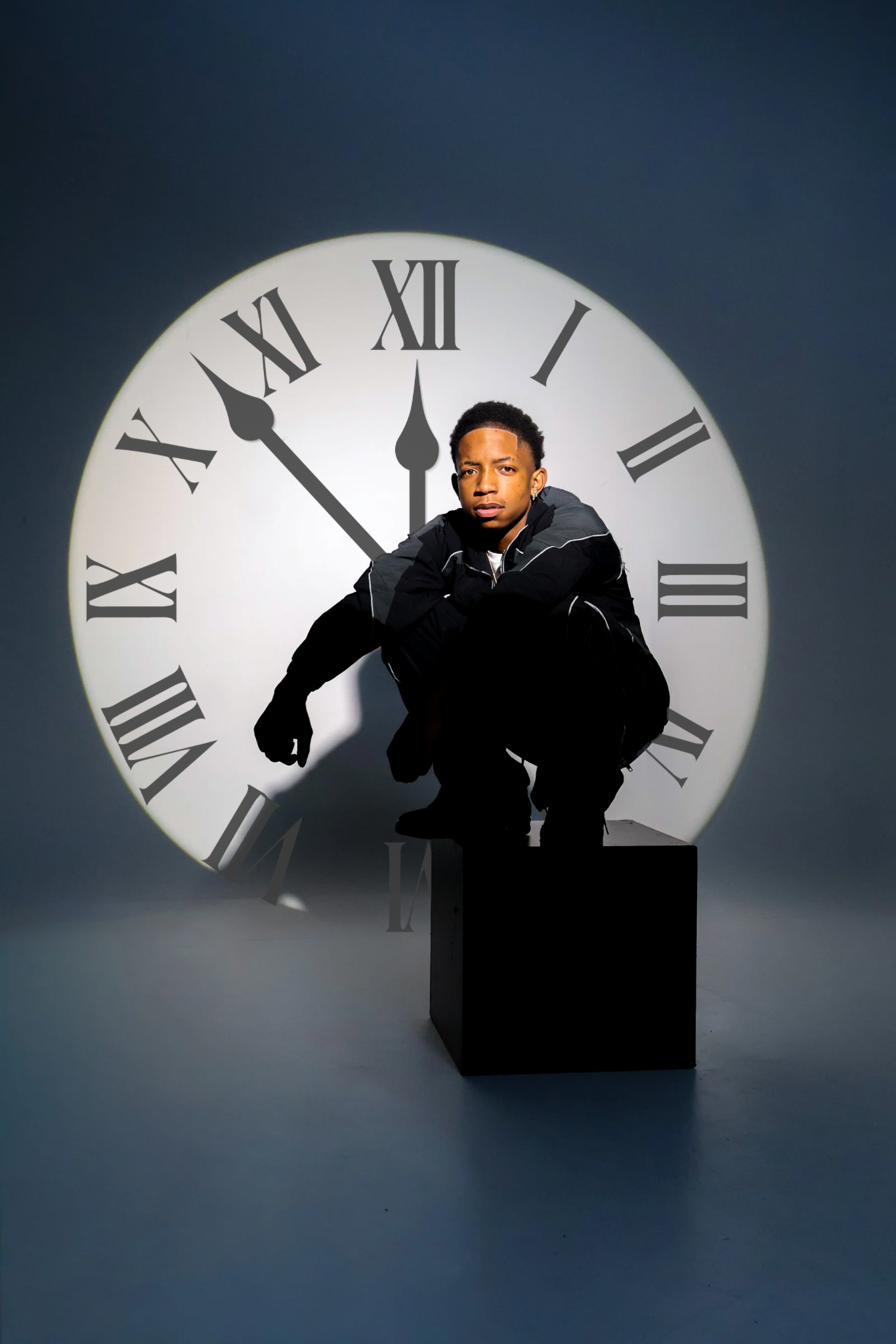 Young man sitting on black cube posing in front of large illuminated clock in dark blue studio.
