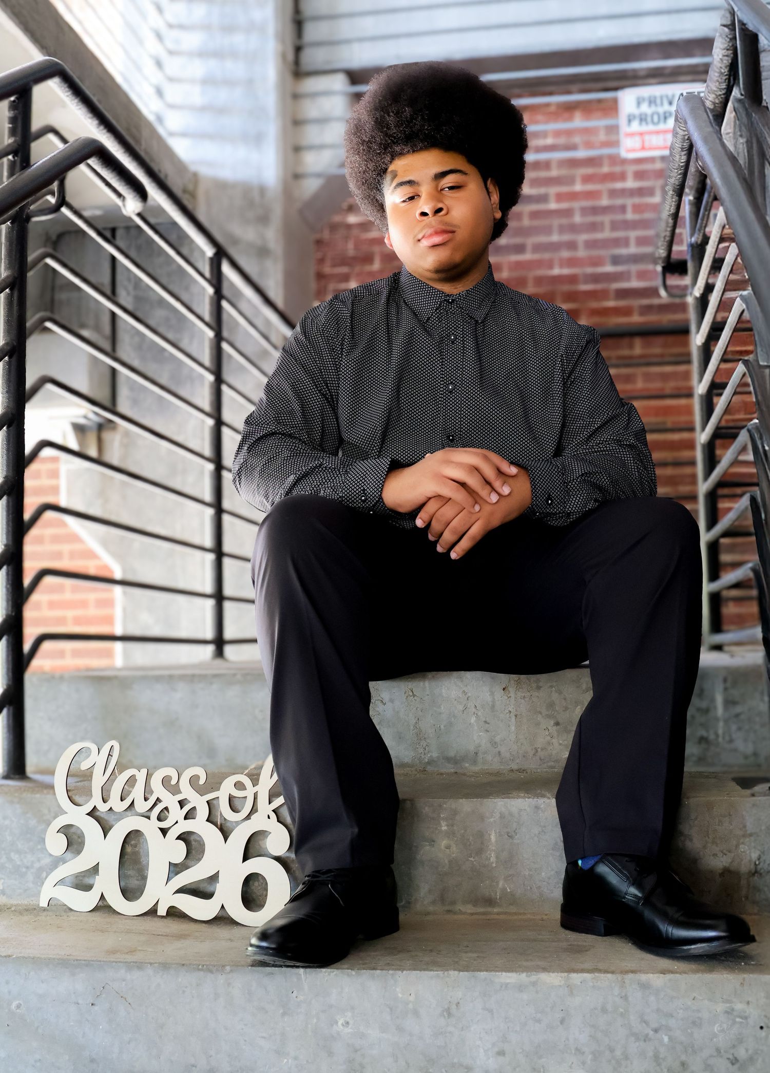 Young man in black outfit sits on concrete steps with Class of 2026 sign for senior portrait photo.