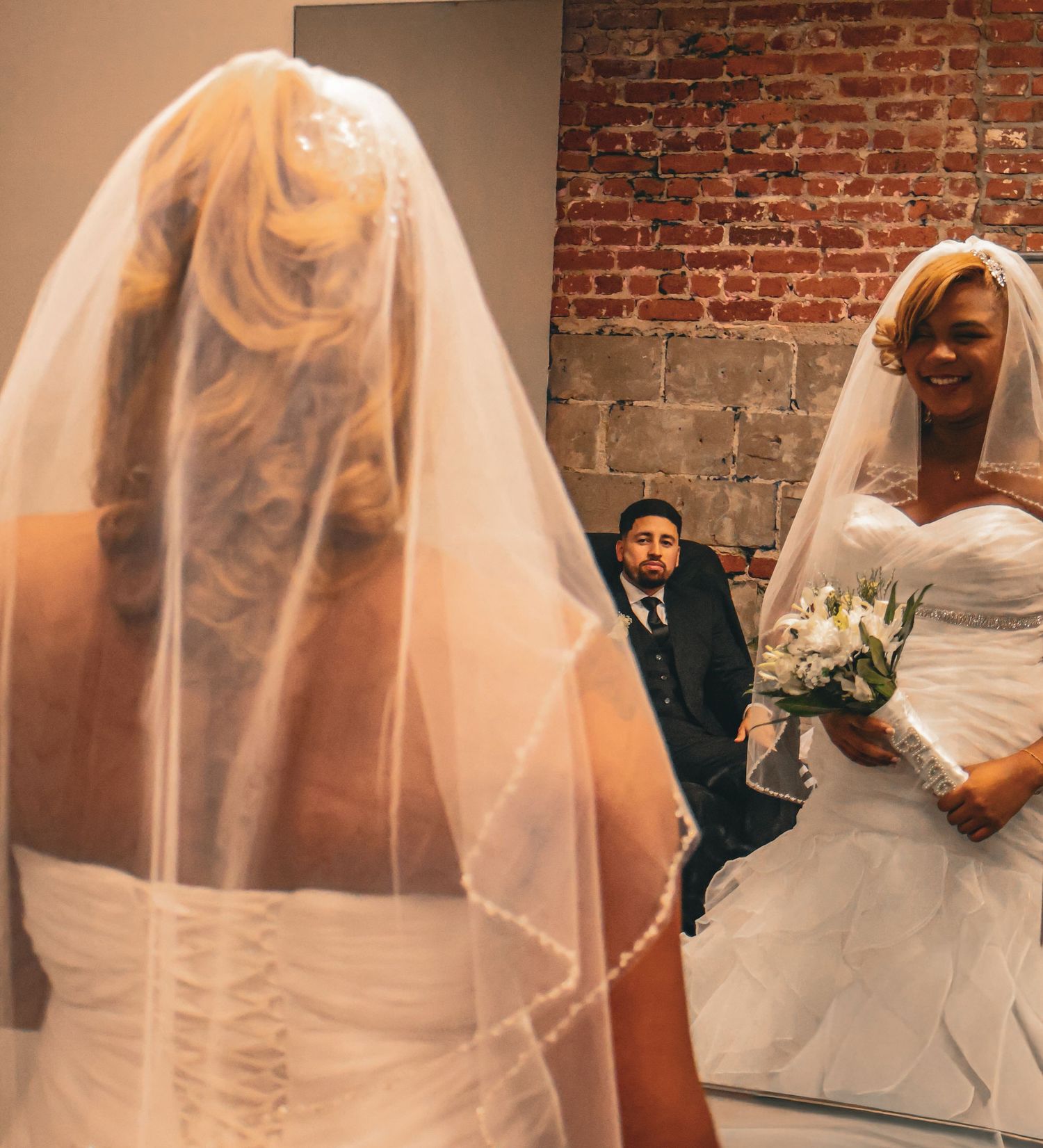 Bride in white gown and veil holds bouquet while groom watches in background near brick wall.