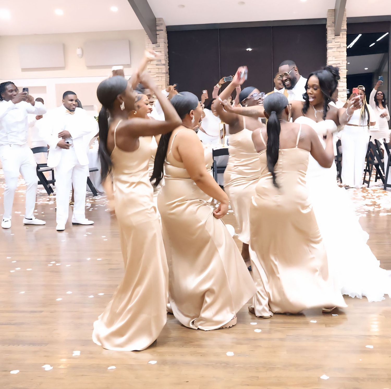 Bridesmaids in champagne satin gowns dancing joyfully at an elegant all-white wedding reception celebration.