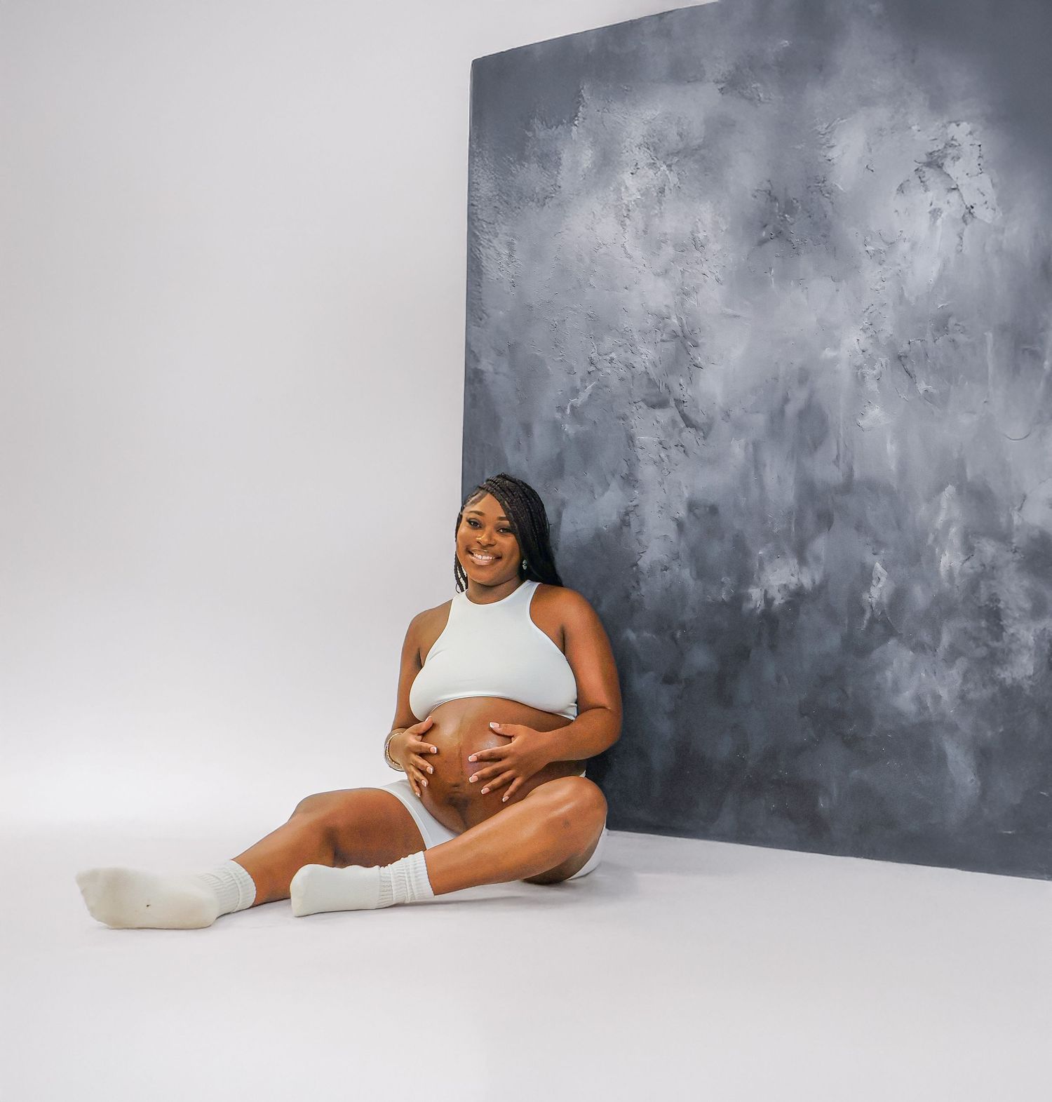 Smiling pregnant woman in white crop top and shorts sits on floor against gray textured backdrop, cradling her baby bump.