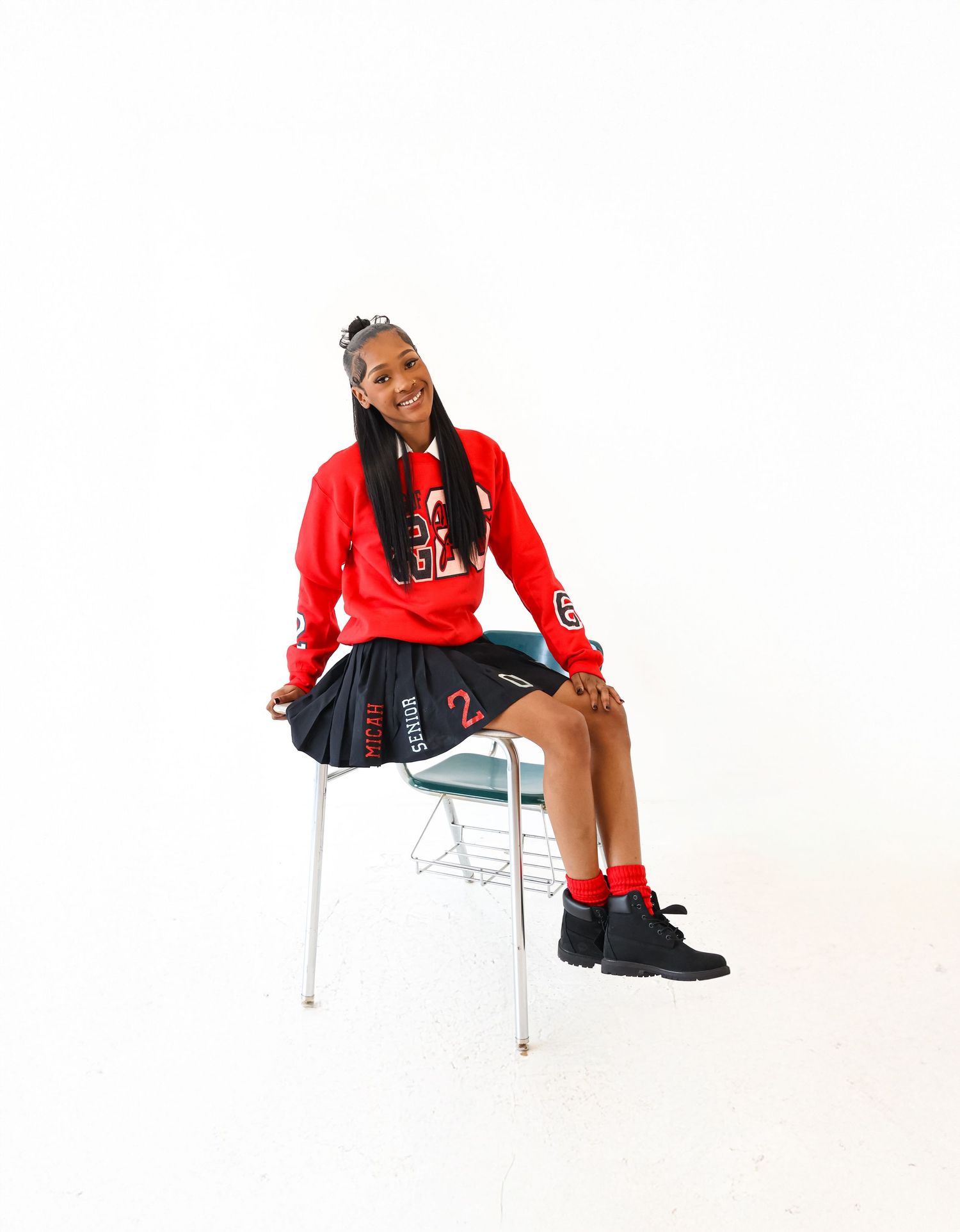 Young girl in red hoodie and pleated skirt sits smiling on a school chair against white background.