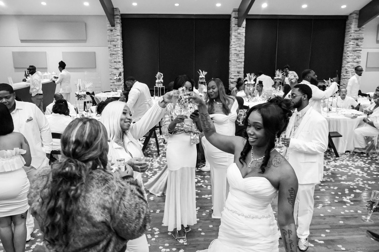 Black and white photo of bride celebrating with guests during lively wedding reception indoors.