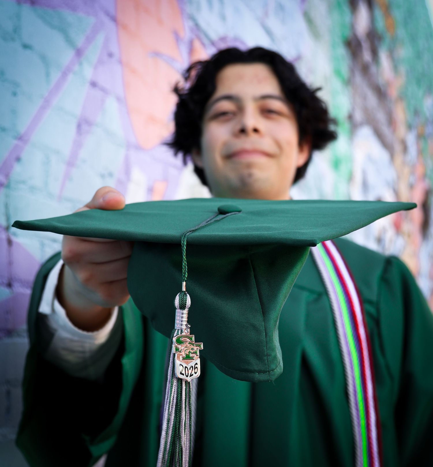 A graduate in green cap and gown holds mortarboard toward camera with tassel and charm visible.