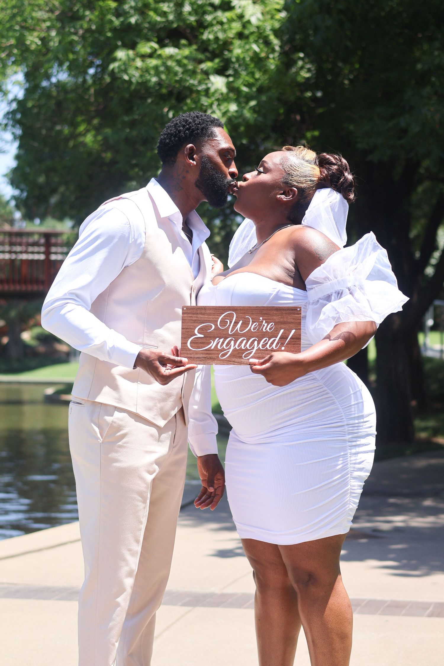 Engaged couple kissing while holding We're Engaged wooden sign in sunny park setting.